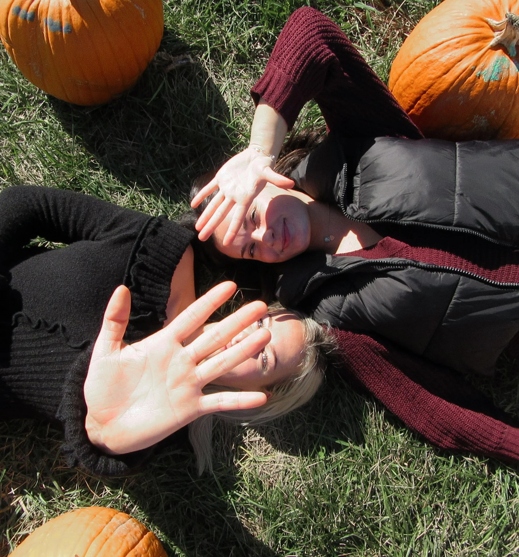 A person lying on grass outdoors surrounded by pumpkins, with one hand covering part of their face and another reaching towards the camera.