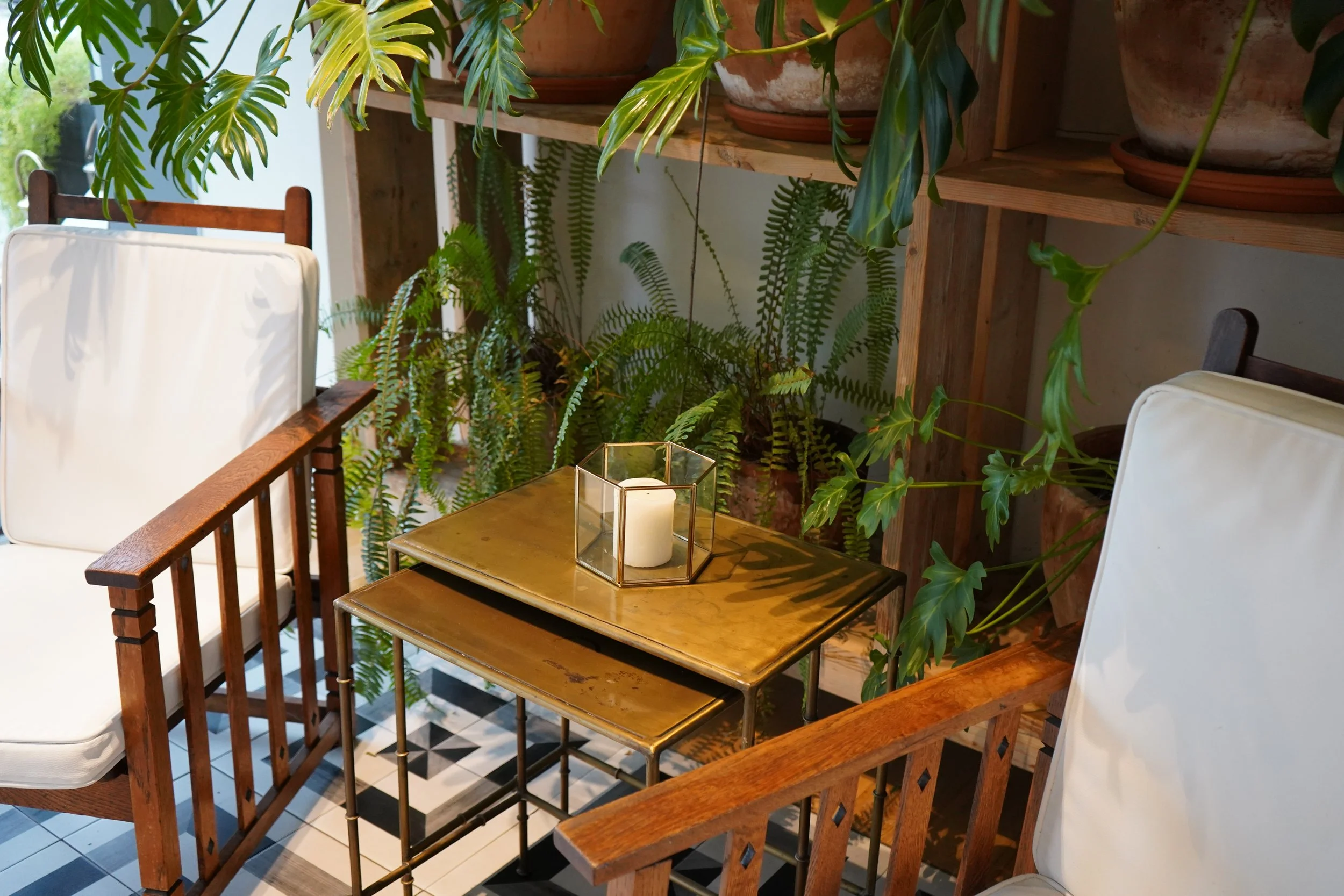 A dining area with a wooden table and matching chairs, surrounded by various green plants in pots and on shelves. A glass candle holder with a white candle is on the table, and the floor features a black and white checkered tile pattern.