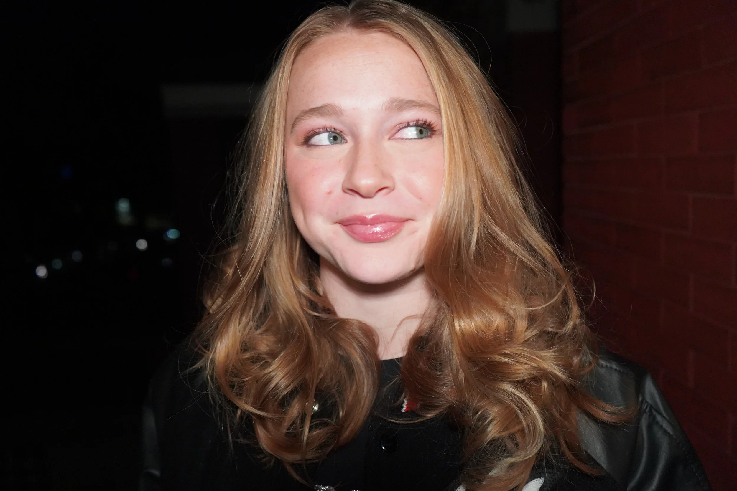 Close-up of a young woman with long, wavy red hair, fair skin, and light makeup, standing outdoors at night with a dark background and faint city lights.
