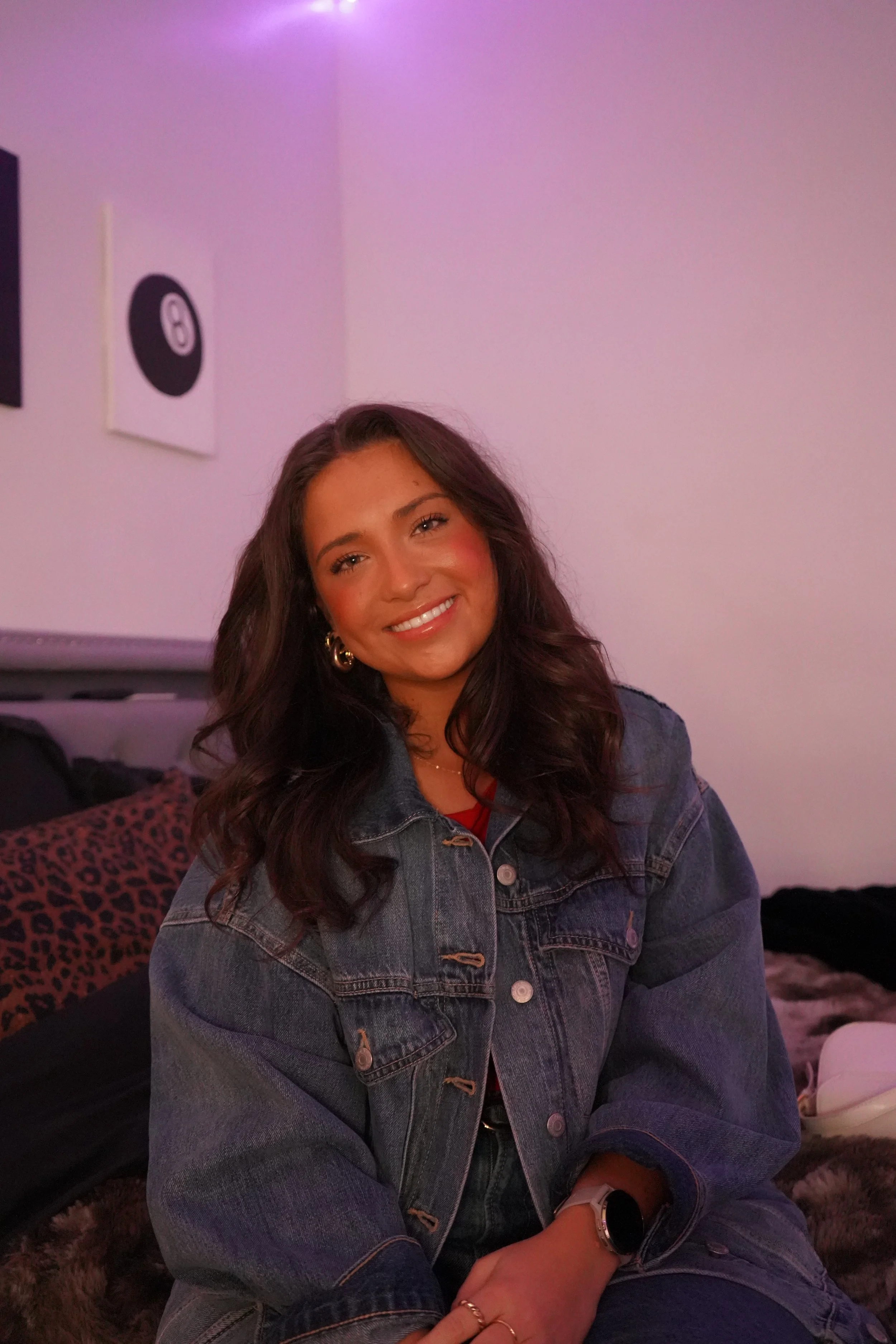 A woman smiling with long wavy hair, wearing a denim jacket and jewelry, sitting in a room with pink/purple lighting, against a wall with art prints of black and white circles and black rectangles.