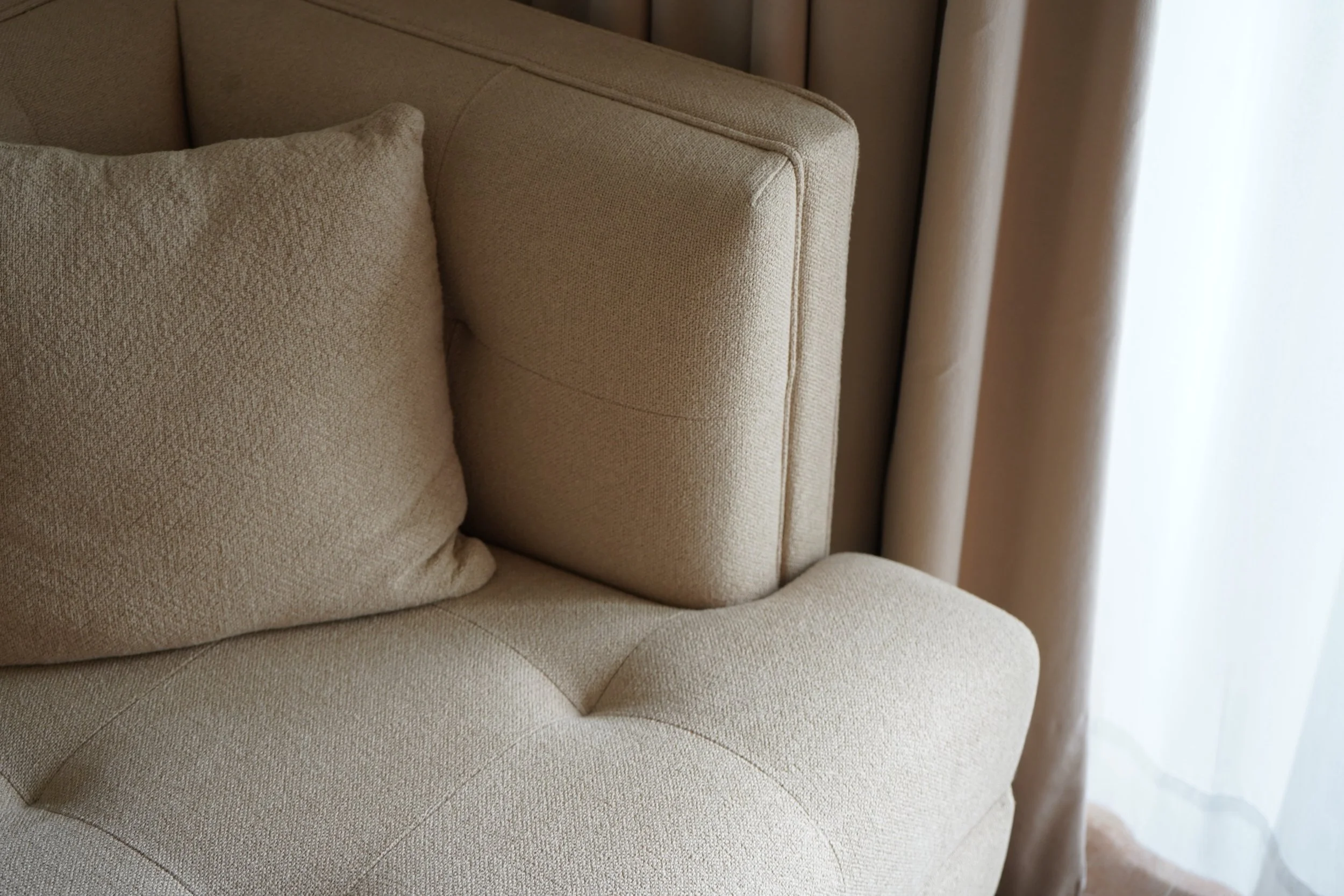 Close-up of a beige upholstered armchair with a cushion and curtain in the background.