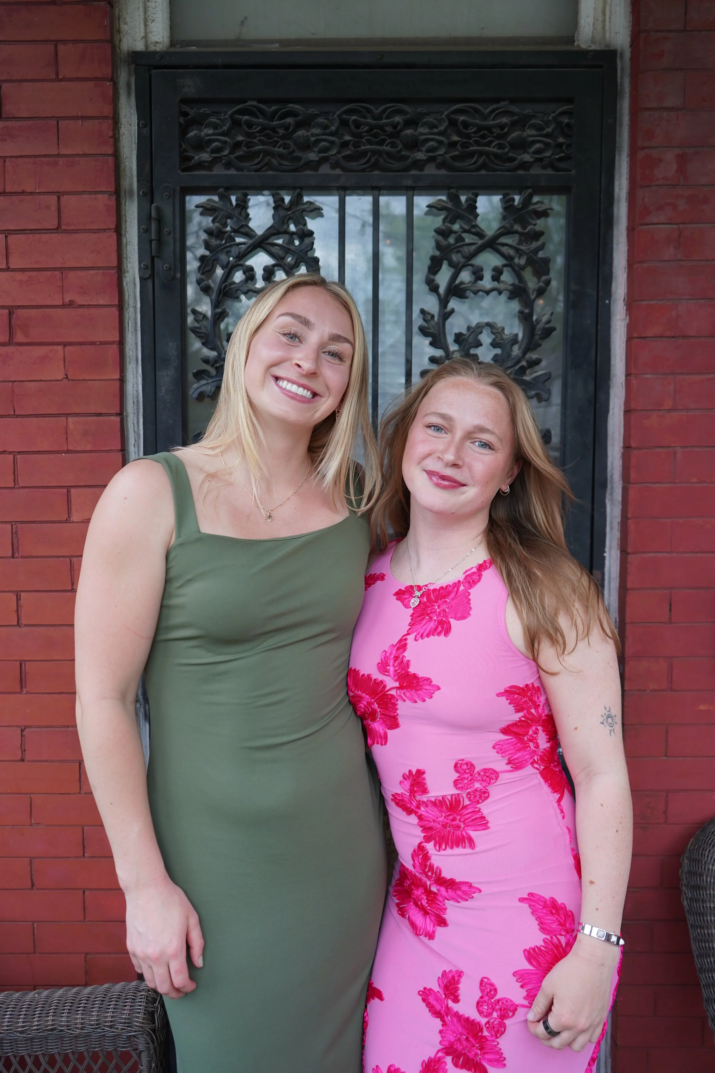 Two women standing in front of a decorative black window on a red brick wall, smiling at the camera.