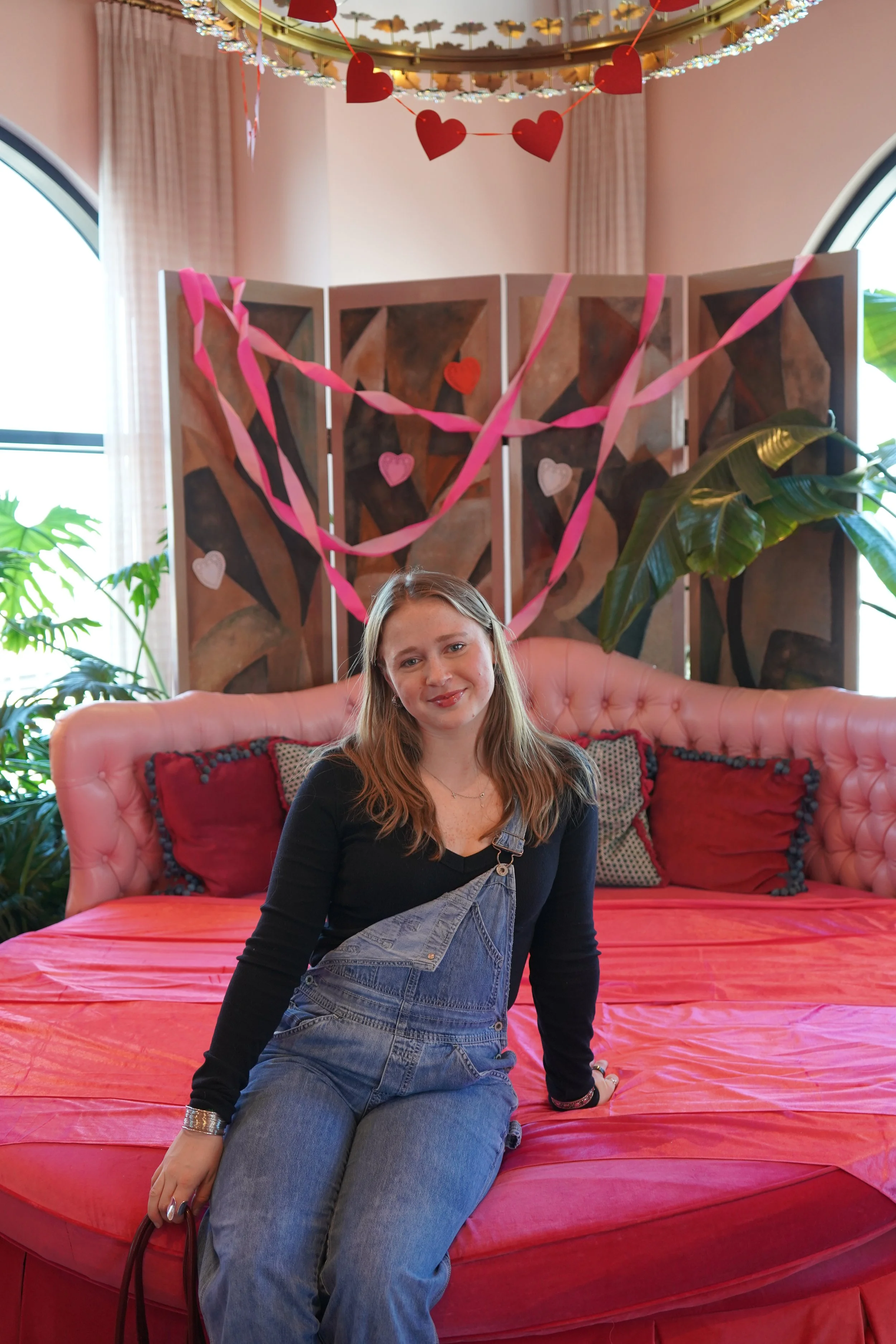 A woman with long hair and a black shirt sitting on a pink couch, with a decorative room with heart-shaped and pink streamers hanging above her, and large green plants flanking the sides of the scene.