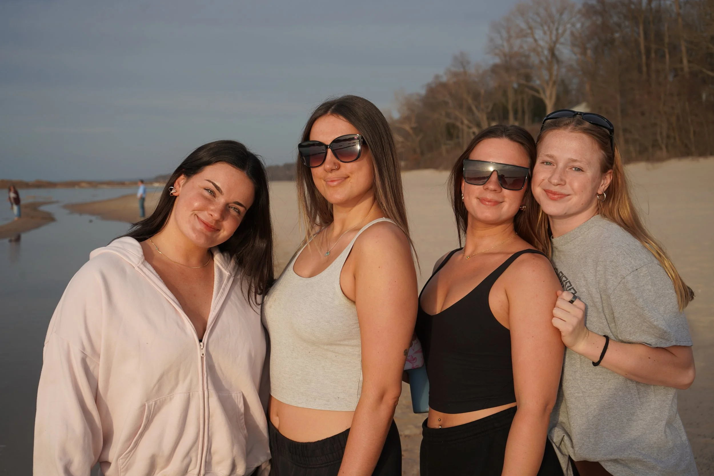Four women standing close together outdoors near a body of water with trees in the background, smiling and wearing casual clothing and sunglasses.