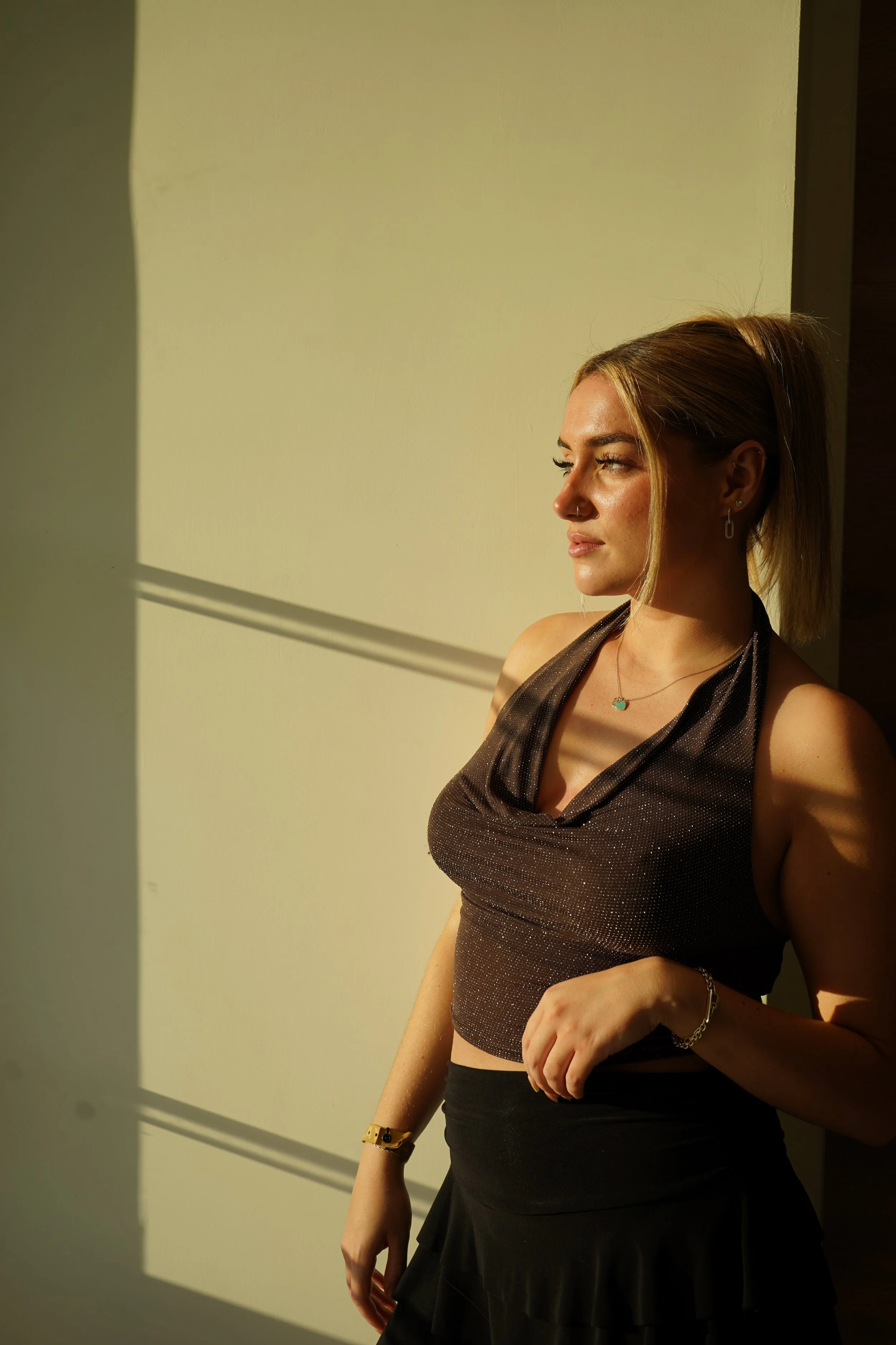 A woman with blonde hair, earrings, and a nose piercing standing in sunlight casting shadows on a yellow wall.