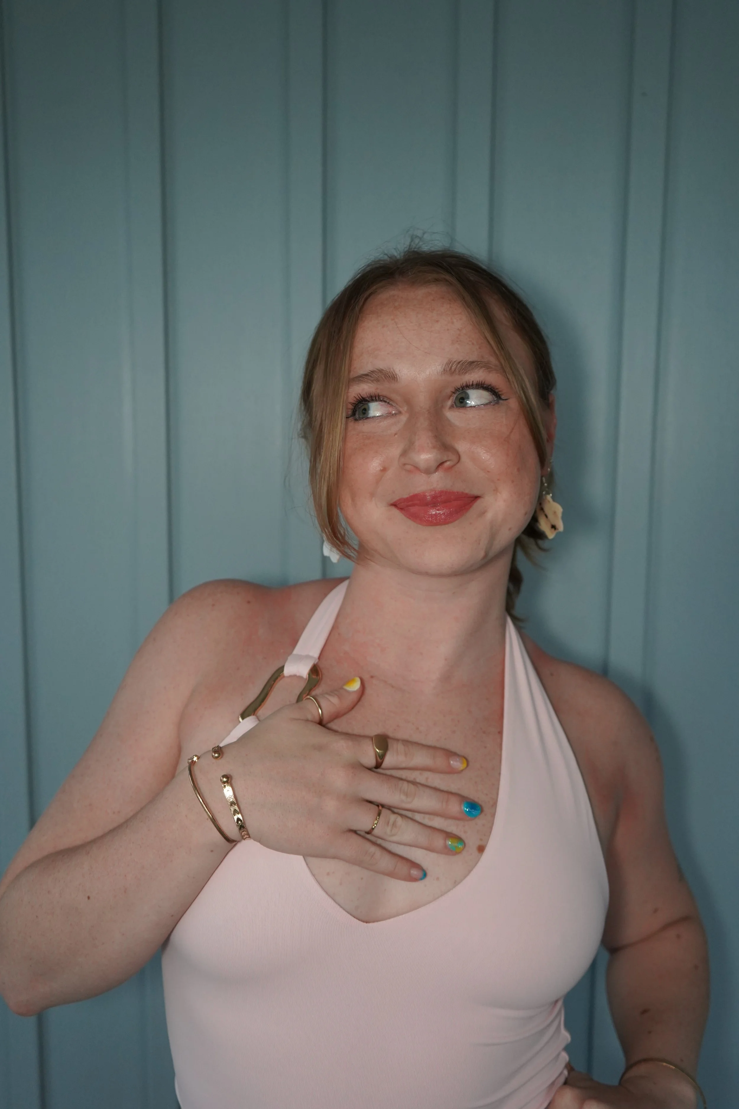 A young woman with red hair, freckles, and blue eyes, smiling and looking to her left. She wears a light pink top and colorful nail polish, with jewelry including bracelets, rings, and earrings. She is standing in front of a blue wooden wall.
