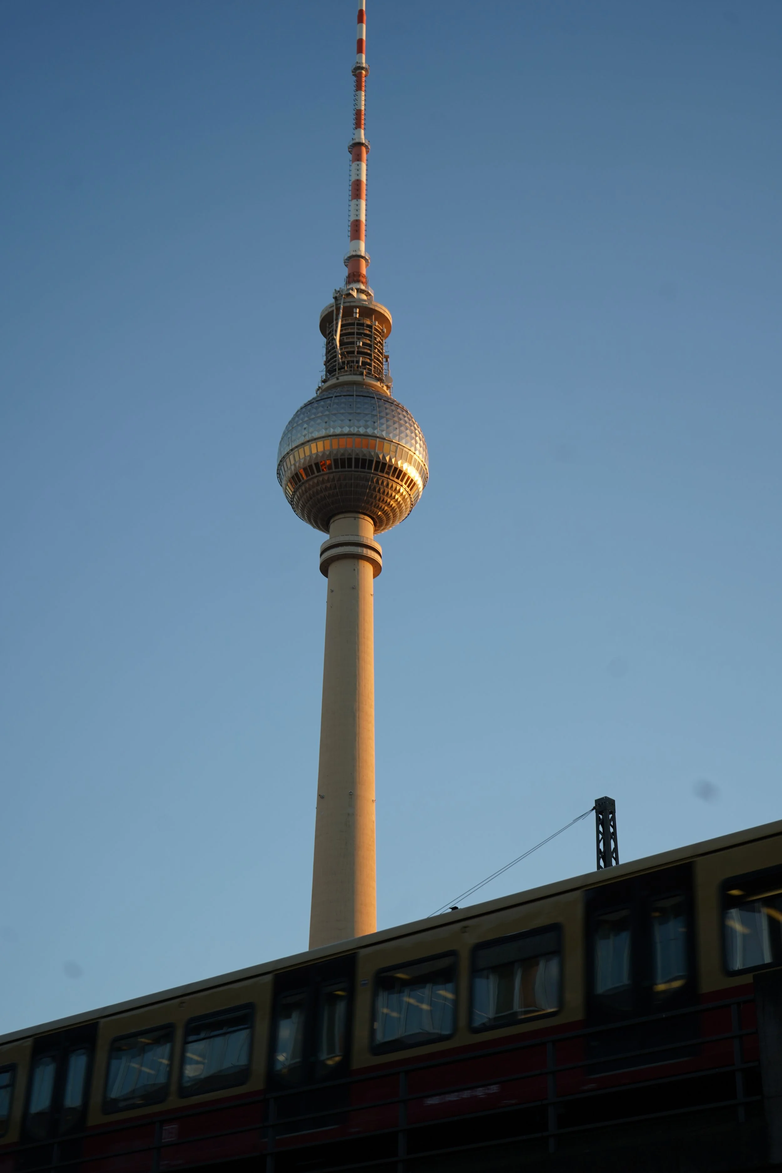 A city observation tower with a spherical section and a long, striped antenna reaching into a clear blue sky, with a train passing on a bridge in the foreground.