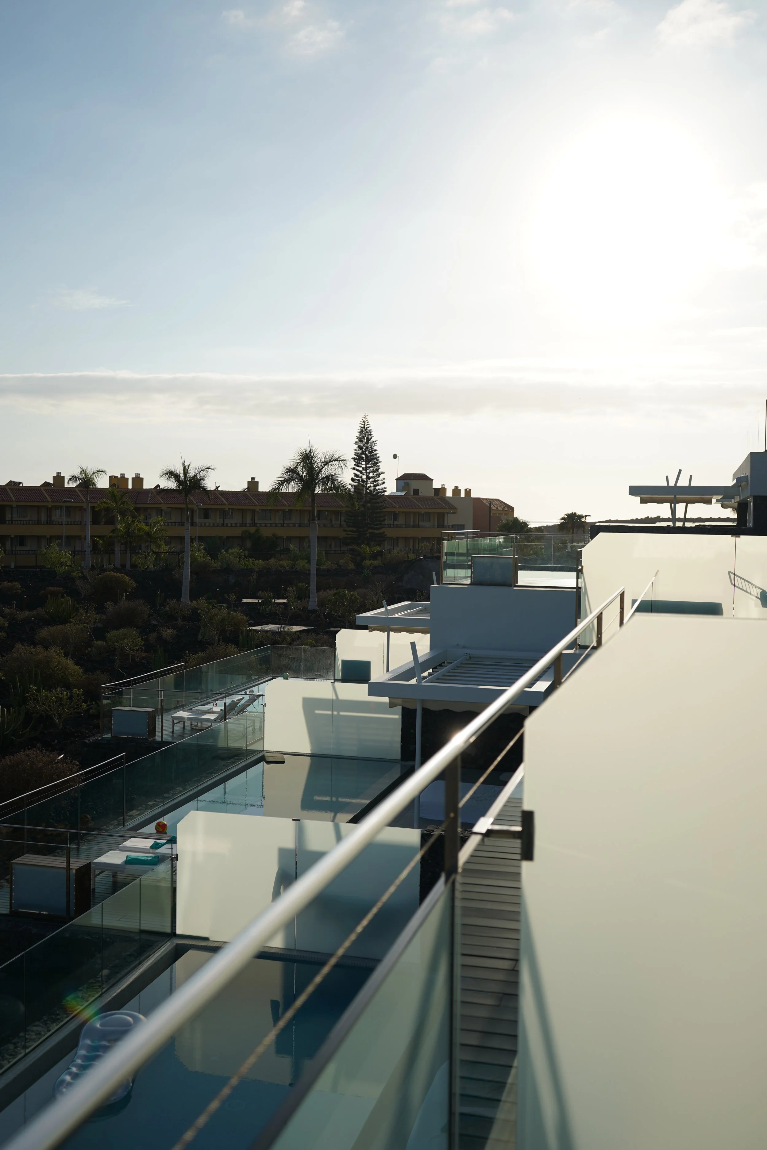 View of modern white building with terraces, glass railings, and outdoor furniture, set against a background of trees, residential buildings, and a bright sky with the sun shining.