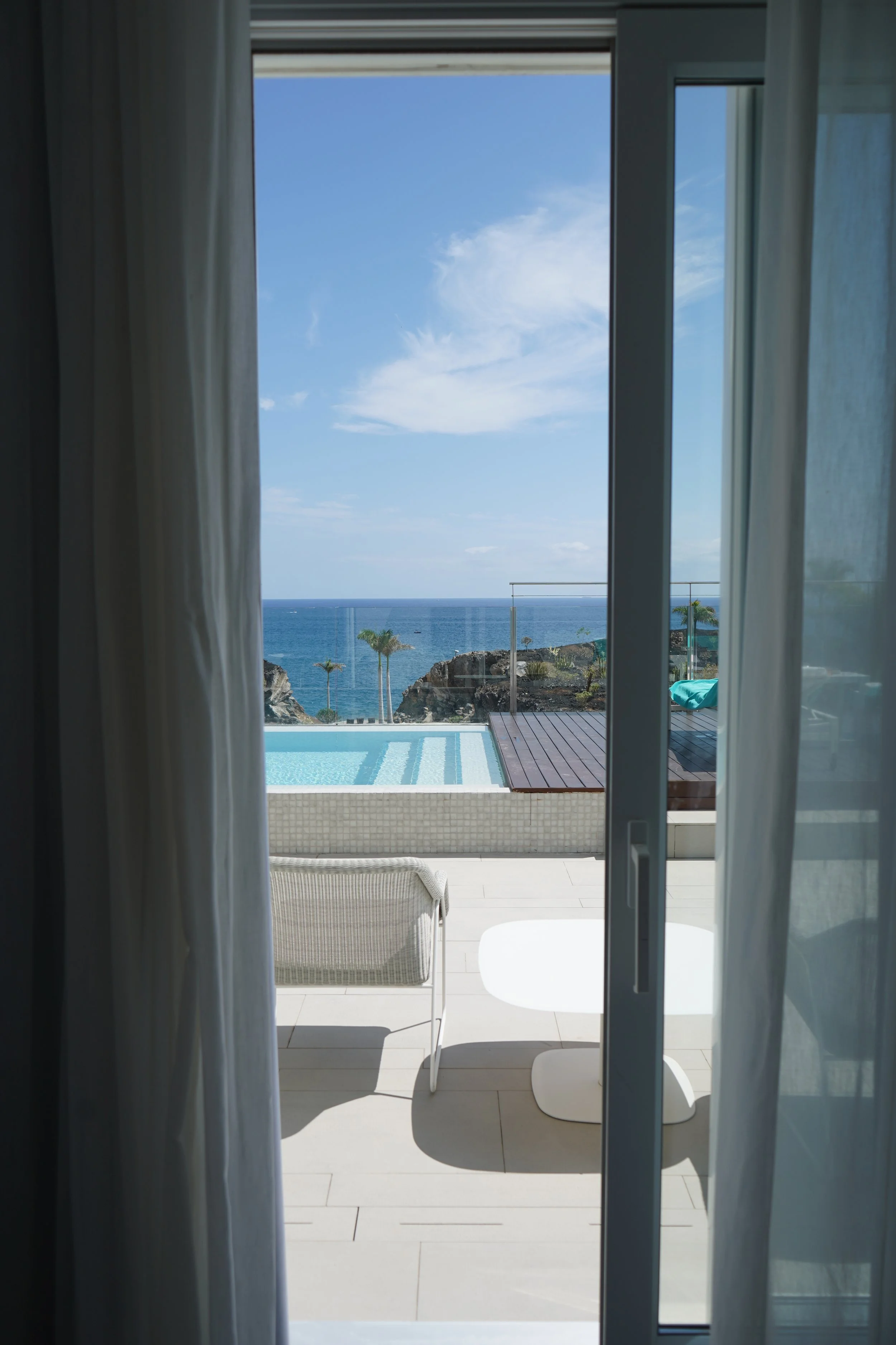View of a pool and ocean through sliding glass doors, with white curtains on either side.