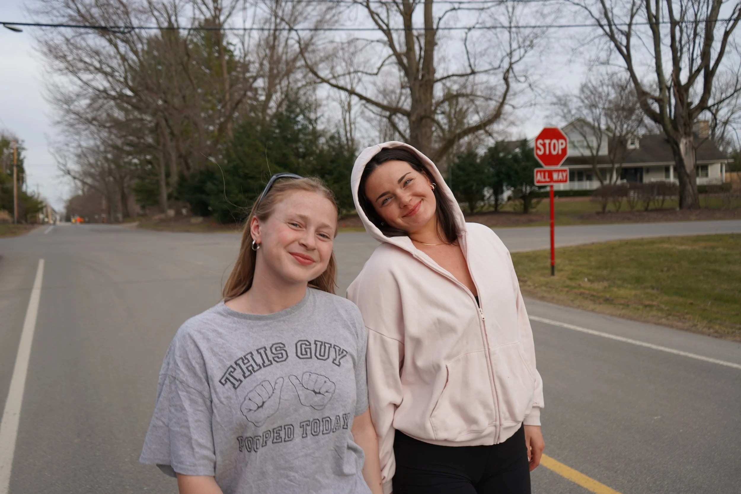 Two smiling young women standing outdoors on a street corner, with a stop sign and houses in the background. One woman has long blonde hair and is wearing a gray t-shirt, and the other has dark hair with a hoodie, wearing a light-colored zip-up jacke