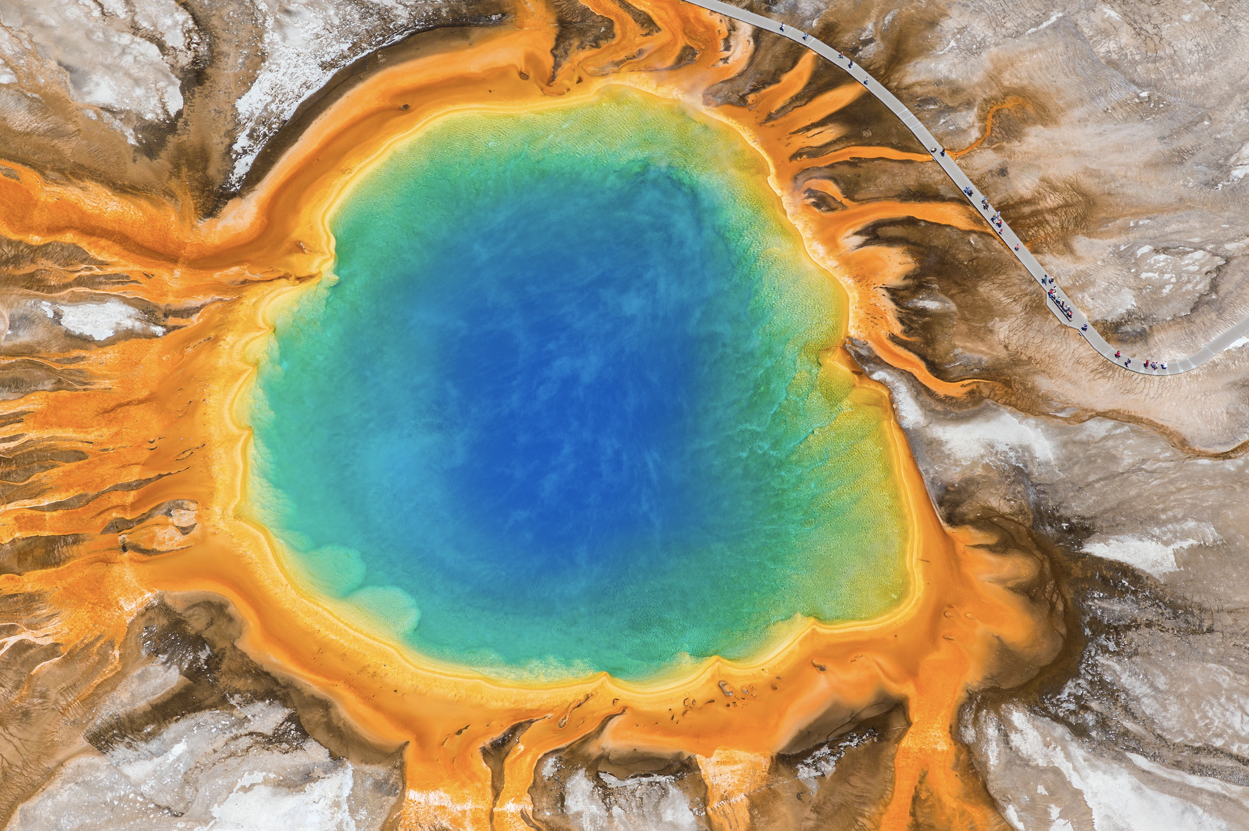 A colorful hot spring with bright blue water in the center, surrounded by vibrant orange and yellow mineral deposits, with a winding boardwalk on the right side.