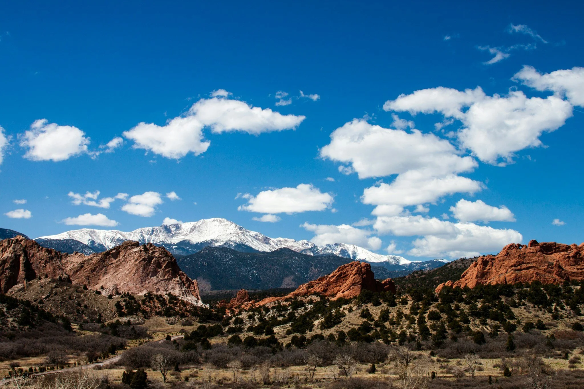 Scenic view of red rock formations in foreground, snow-capped mountains in background, and a bright blue sky with scattered white clouds.