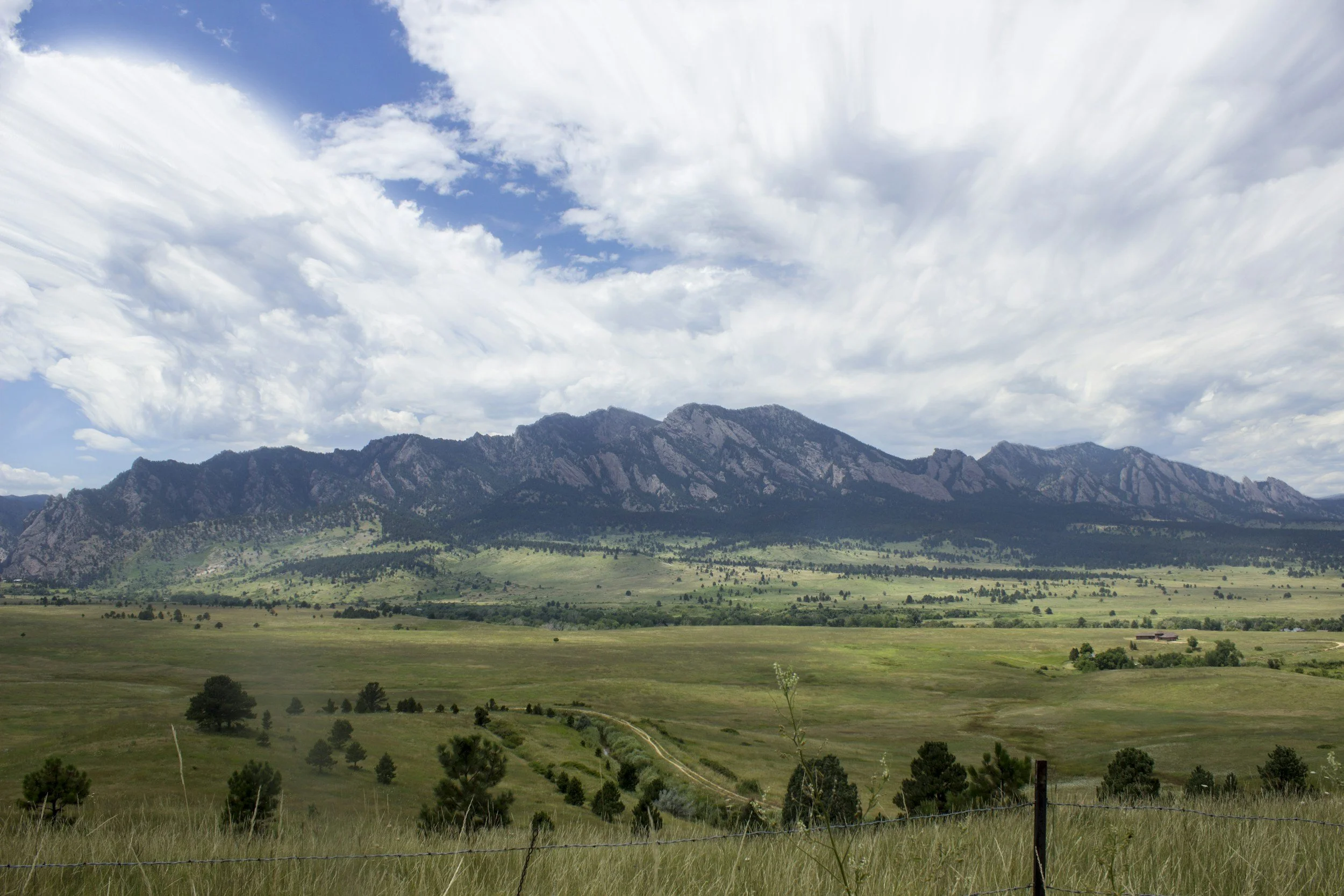 A wide view of a grassy plain with scattered trees, a dirt path, and a mountain range in the background under a partly cloudy sky.
