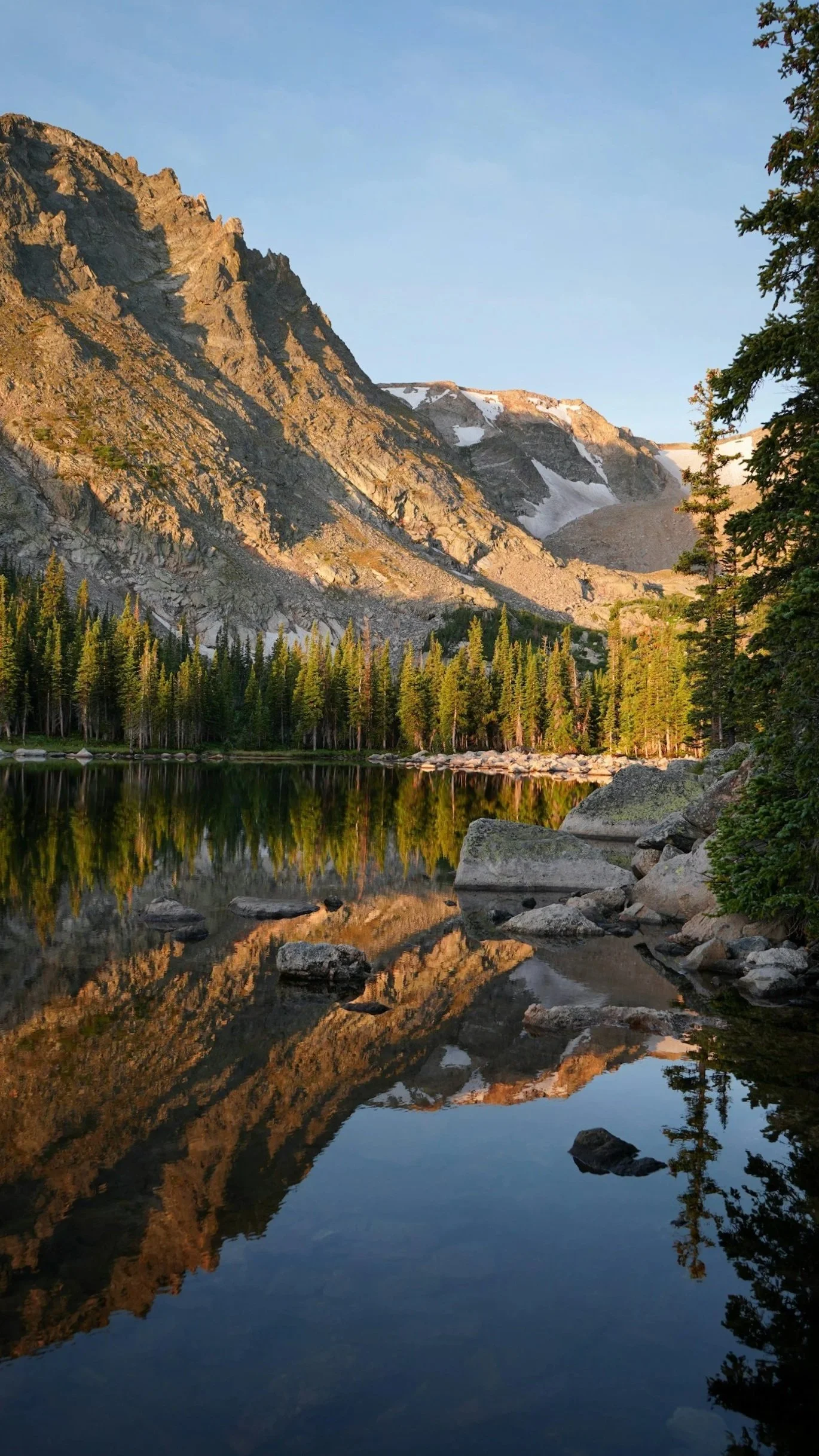 Mountain lake landscape with rocky mountains, pine trees, and their reflection in the calm water during sunset.