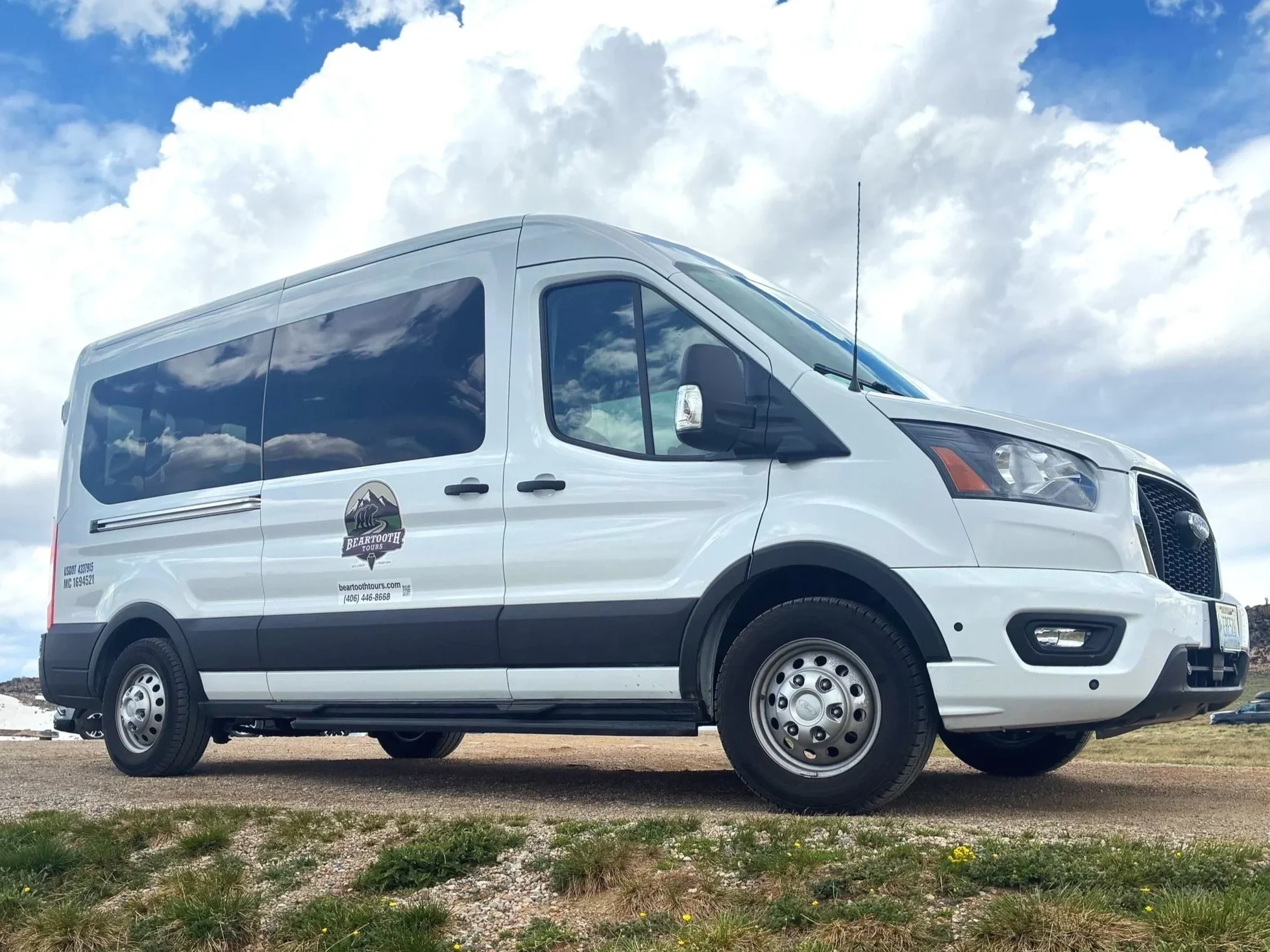 White touring van with tinted windows, parked on gravel ground under a partly cloudy sky, with a logo of mountains and the text ‘Bear Tooth Tours’ on the side.