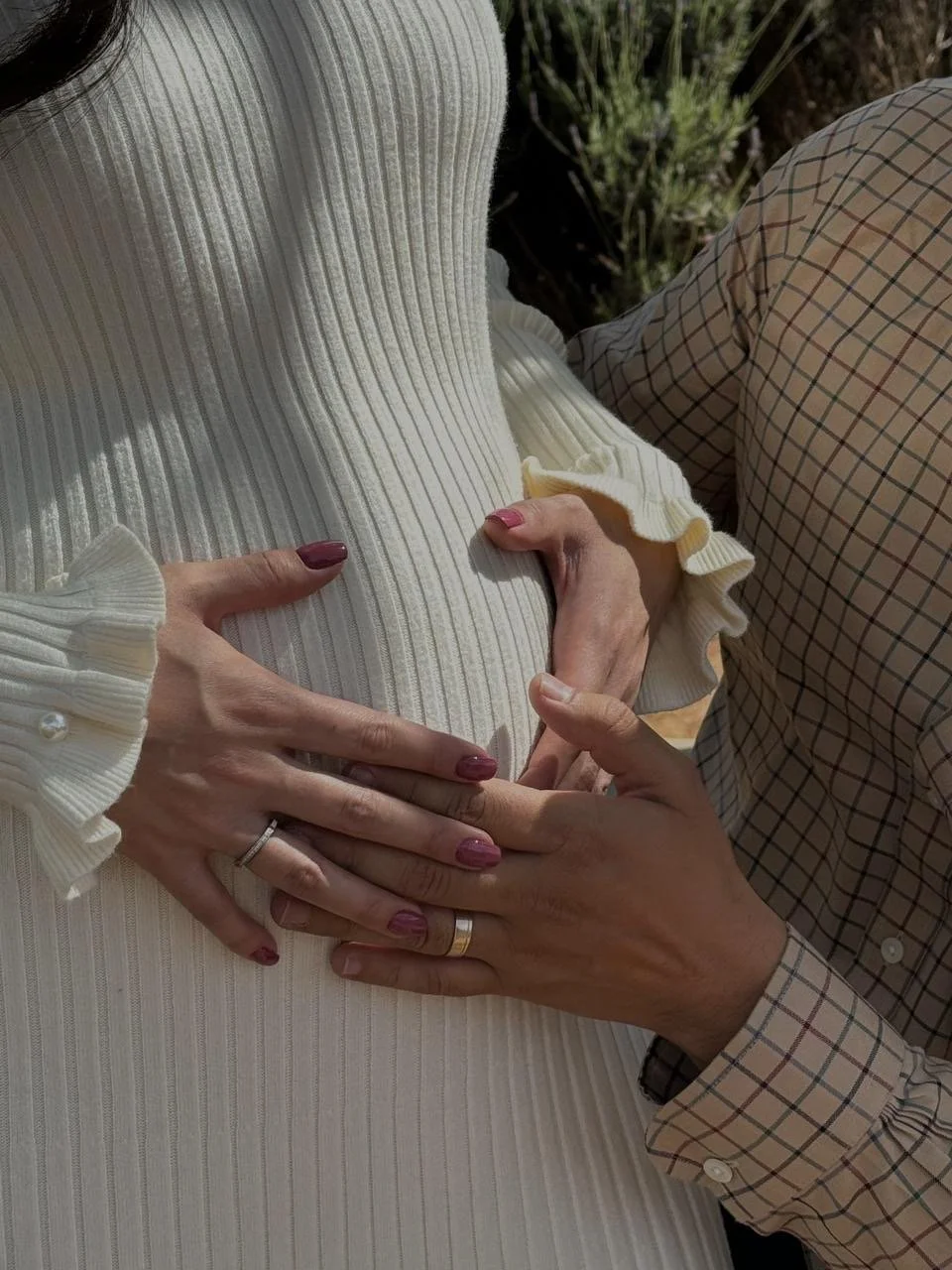 Close-up of two people holding hands on a person's pregnant belly, with one hand placed on top and the other underneath, both wearing rings, and the woman wearing a cream-colored ribbed sweater.