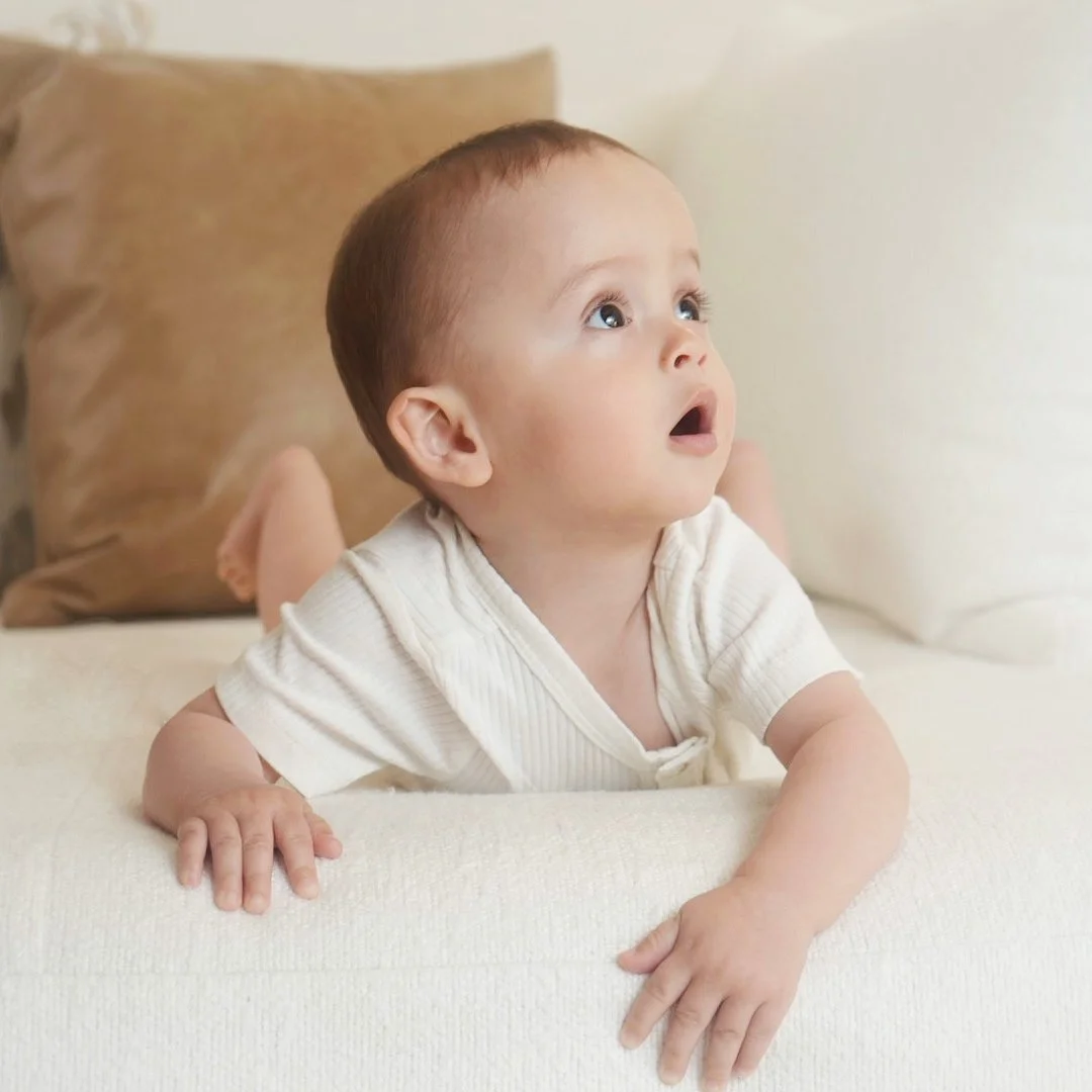A young toddler with light skin and short brown hair lying on their stomach on a bed with beige bedding, looking up with an attentive expression.