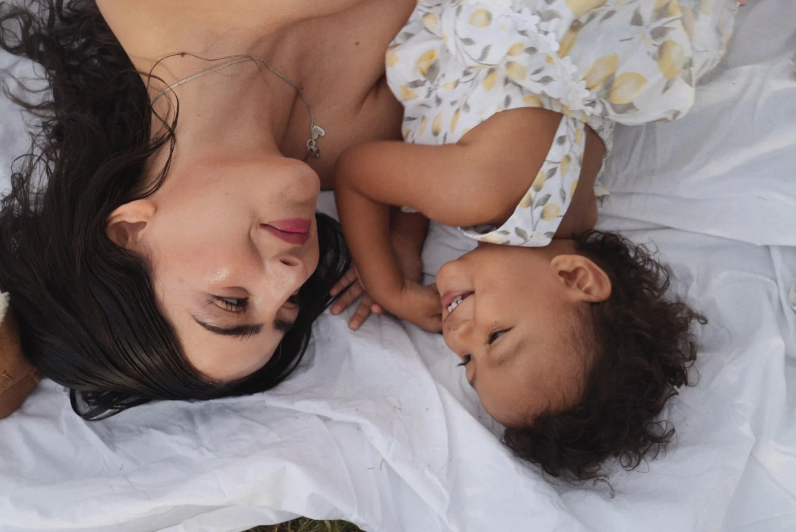 A woman and a young girl lying on a bed, facing each other, smiling and looking into each other's eyes, with white sheets around them.