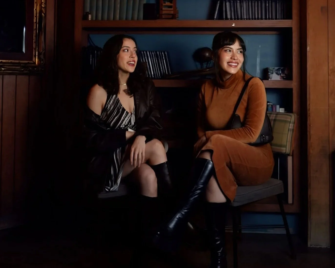 Two women sitting on a bench, smiling and talking in a cozy room with wooden shelves and blue walls.