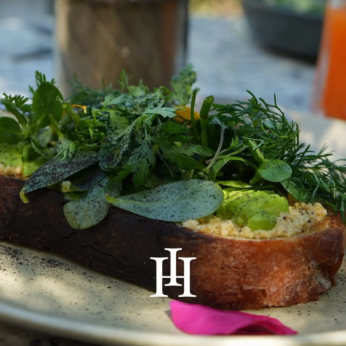 Close-up of an open-faced toast topped with fresh green herbs and vegetables, served on a beige plate with a pink petal and a logo.