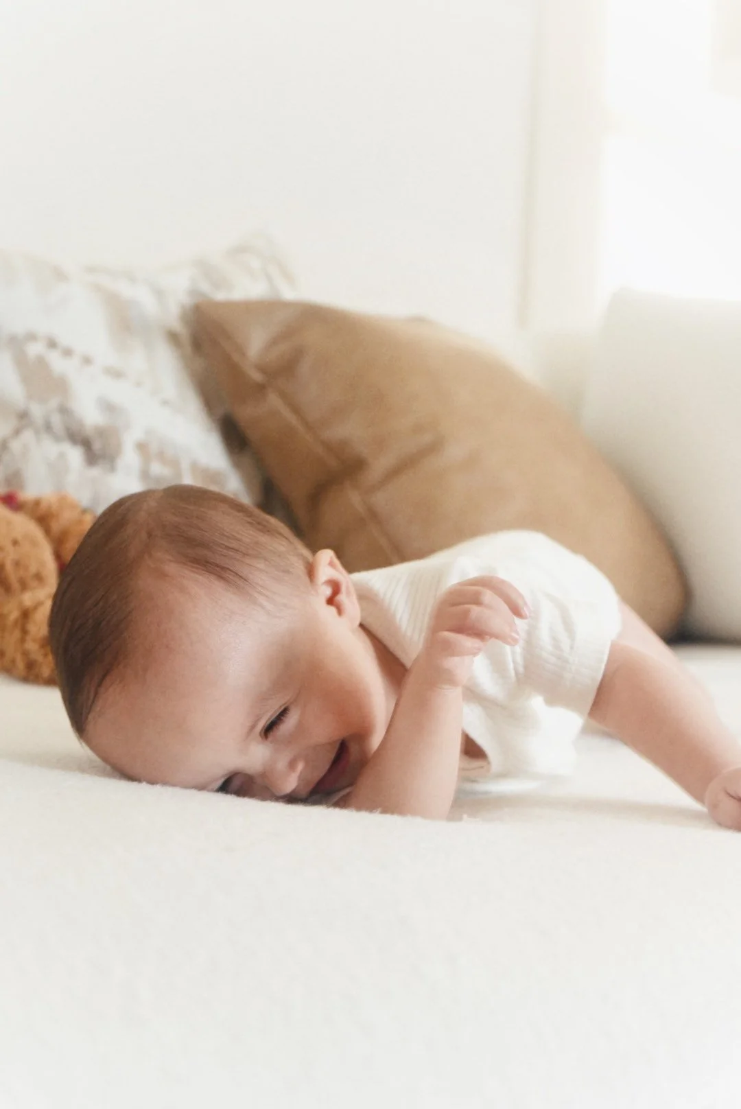 A baby lying on a bed, smiling, with toys and pillows in the background.