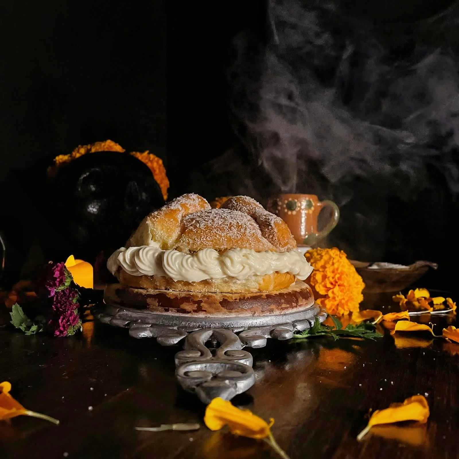 Traditional Mexican three-layered cake topped with whipped cream, dusted with powdered sugar, and garnished with a tamale, with marigold flowers surrounding it, against a dark background.