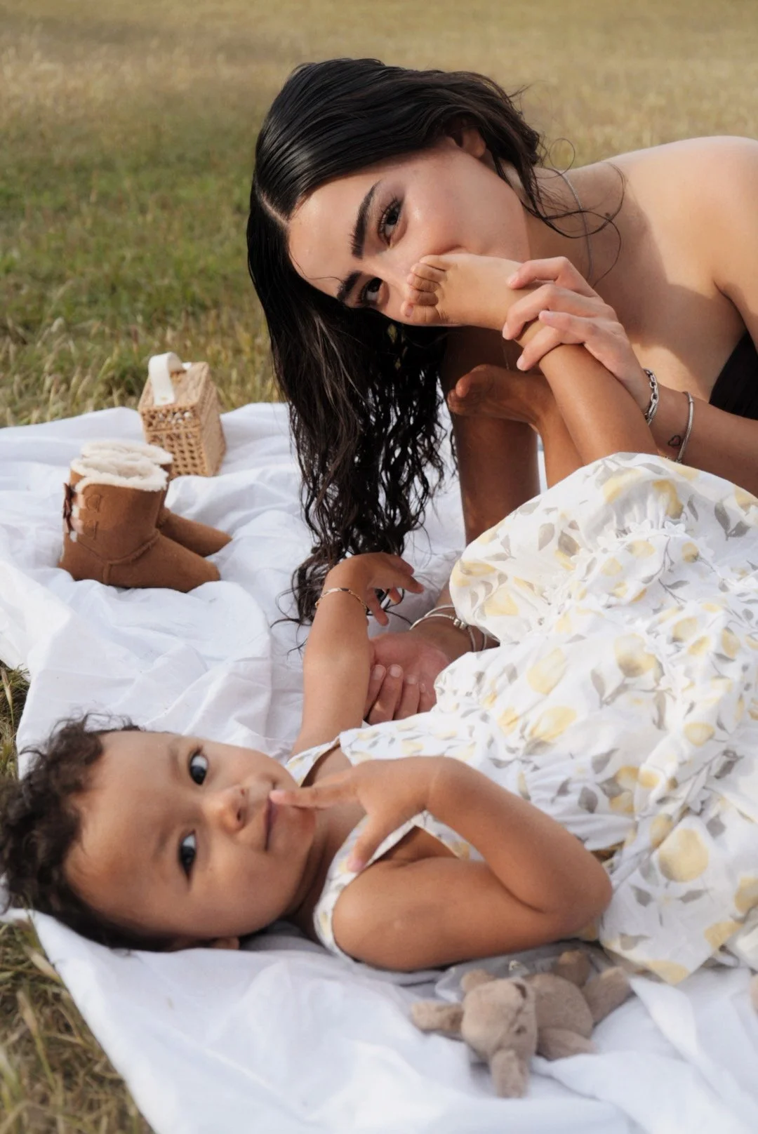 A woman and a young girl lying on a white blanket outdoors on grass. The woman is holding the girl's hand and gently touching her face. The girl is lying on her back, smiling, with her thumb near her mouth. There are two small brown boots and a tissu