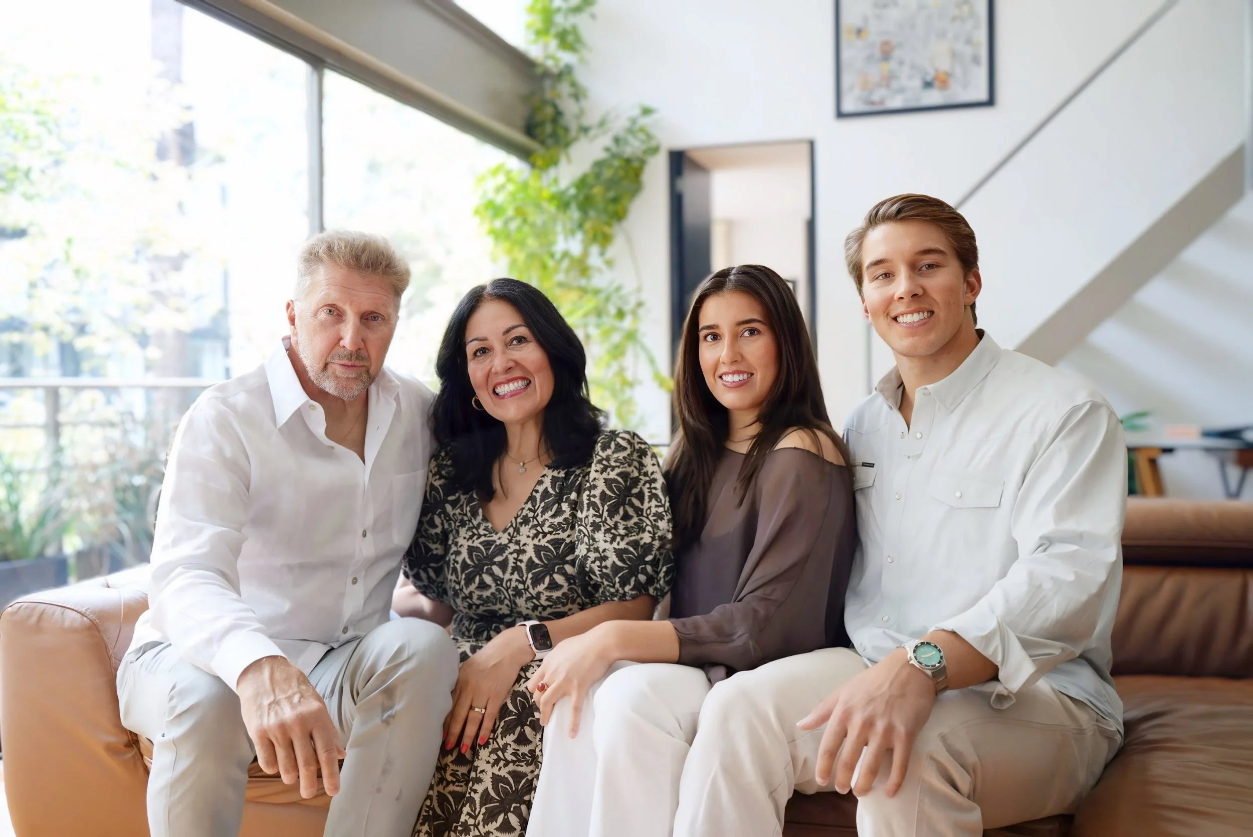 Group of five people sitting on a beige couch in a bright room with large windows and green plants.