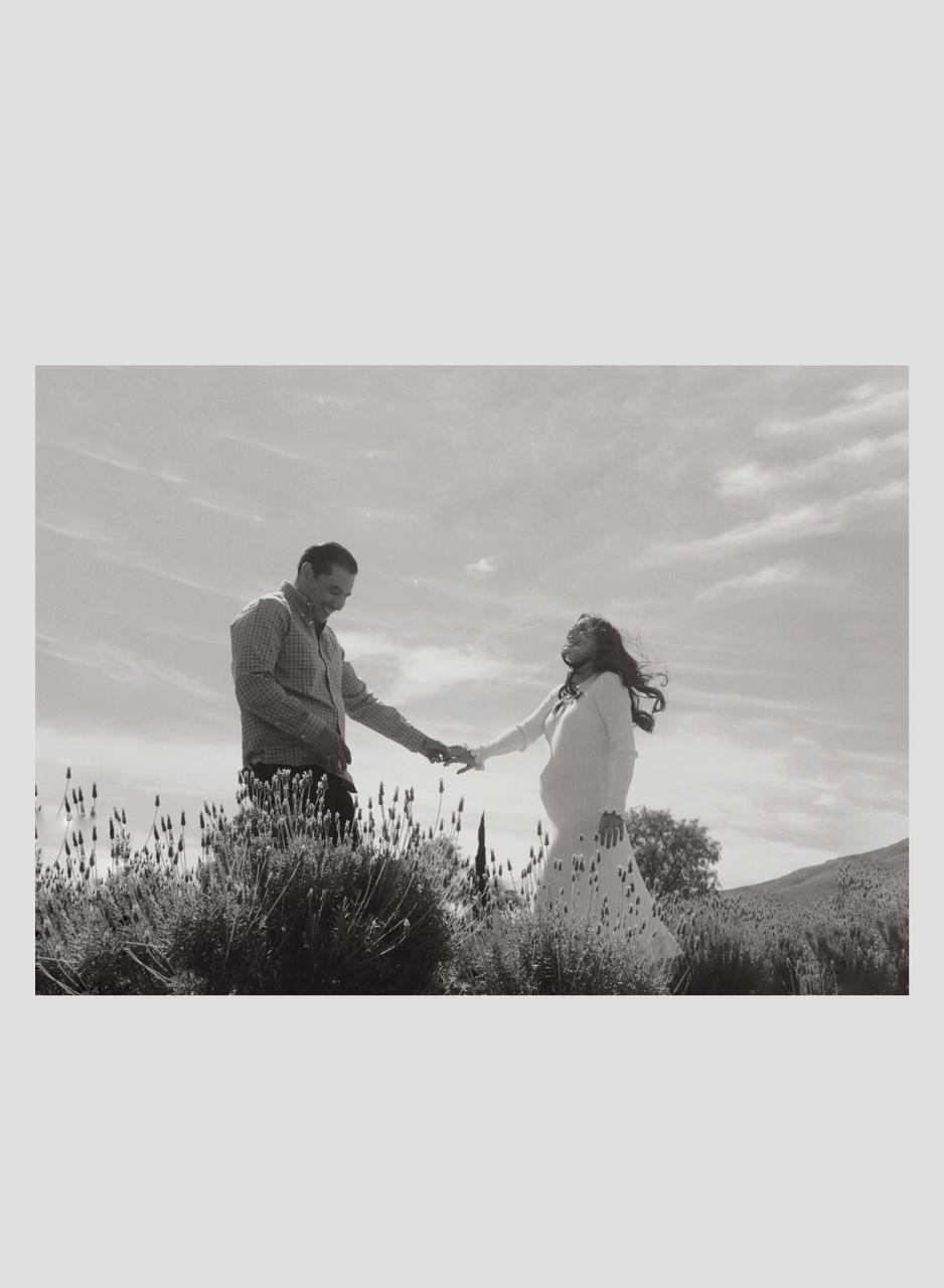 A black and white photo of a couple standing outdoors on a hill, holding hands and facing each other, with flowers and a scenic sky in the background.