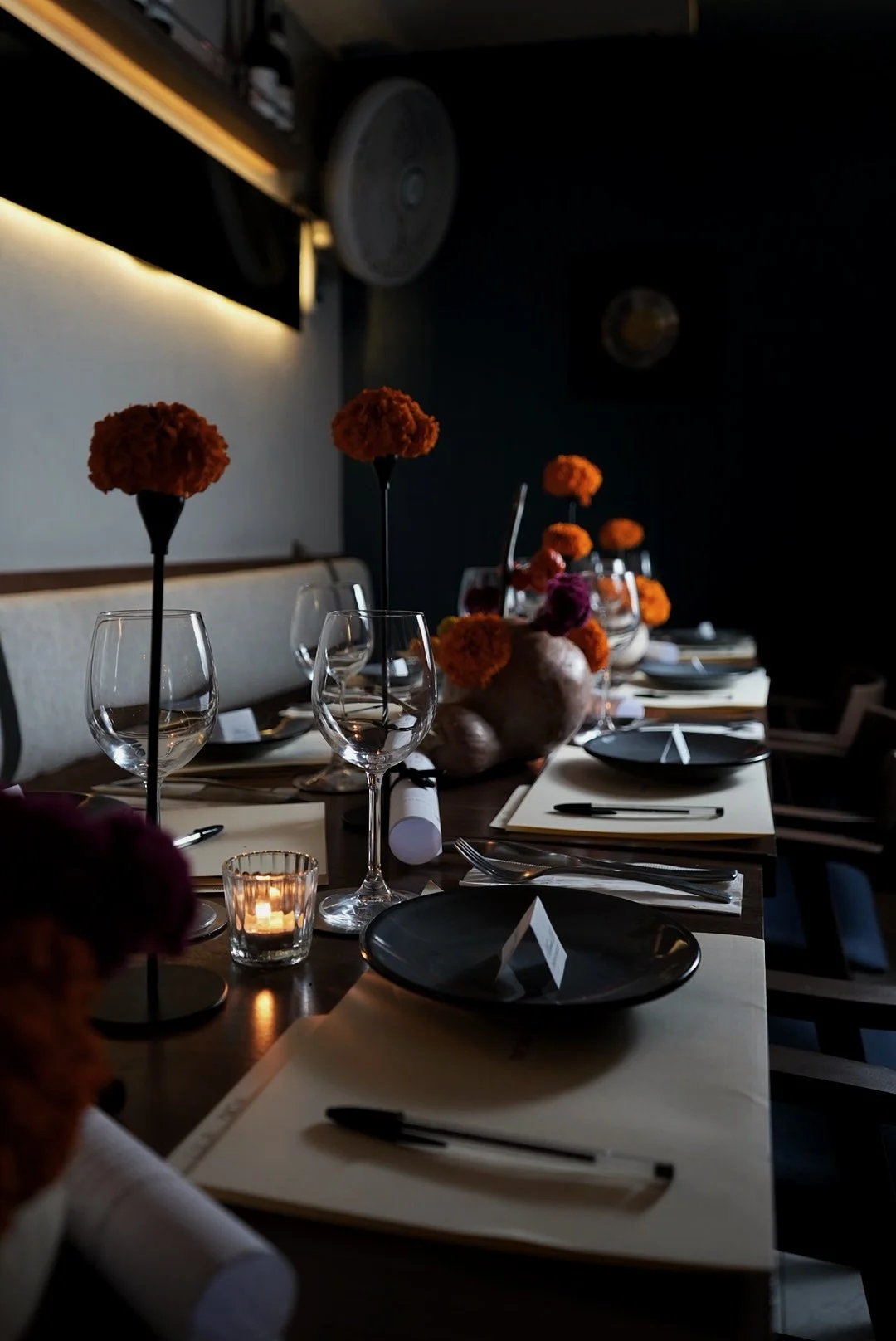 A dimly lit dining table with black plates, glasses, and floral centerpieces with orange and purple flowers, set for a formal meal.