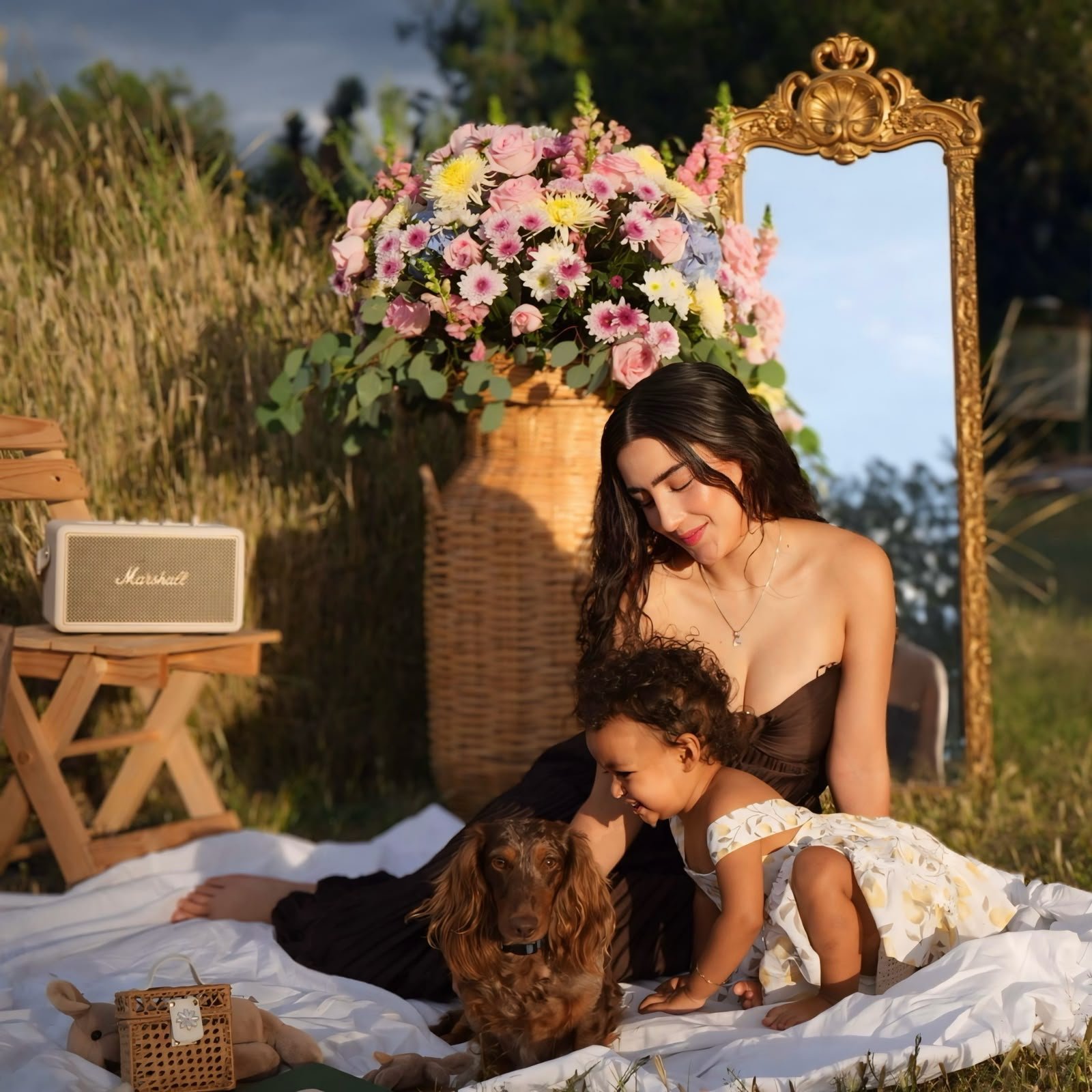 A woman and a young girl are sitting on a white blanket outdoors, playing with a brown dog. Behind them, there is a large flower arrangement in a basket, a gold ornate mirror, and a small speaker. The setting suggests a picnic or outdoor gathering du