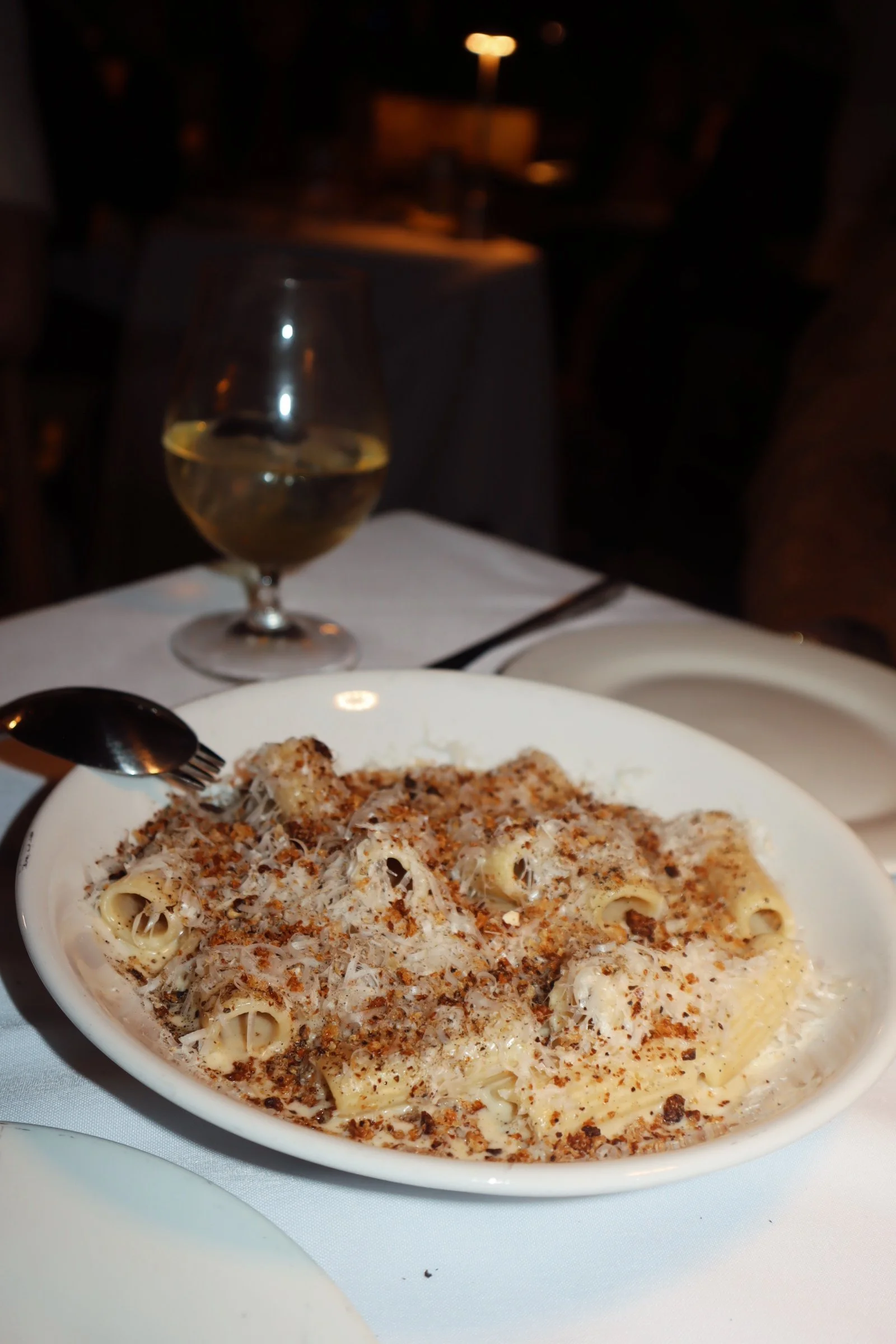 A bowl of creamy pasta topped with grated cheese and breadcrumbs, served on a white table with a spoon, with a glass of white wine and a dark background at a restaurant.