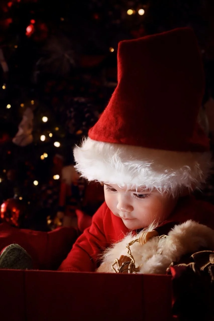 A young child wearing a Santa hat, sitting in front of a decorated Christmas tree, looking at a gift or ornament.