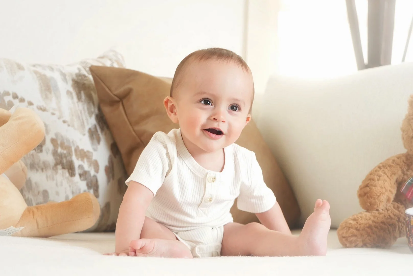 A baby sitting on a couch surrounded by stuffed animals, smiling happily.