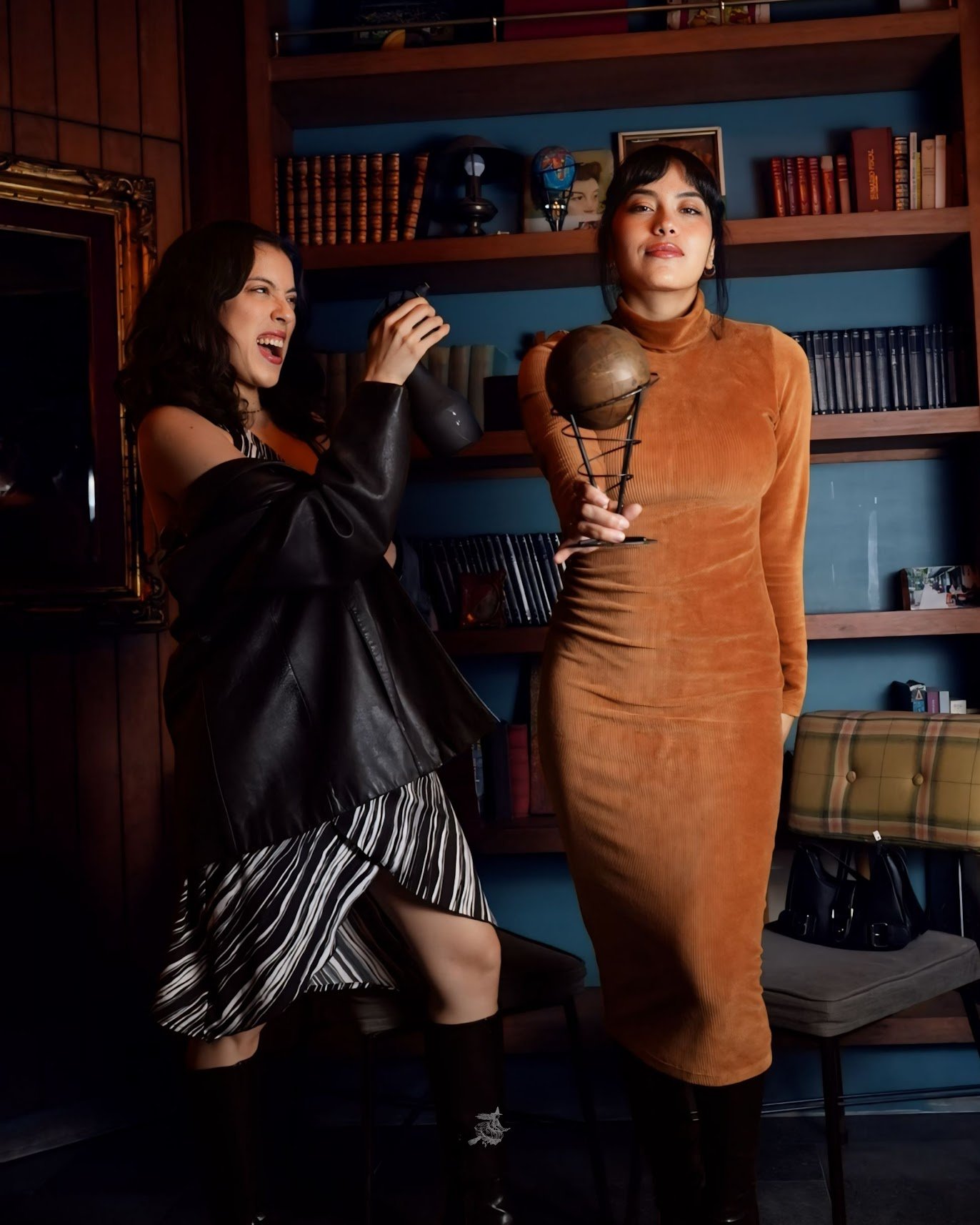 Two women with dark hair posing indoors in front of a bookshelf. The woman on the left is smiling and holding a black object, wearing a striped dress and black jacket. The woman on the right is standing with a slight smile, wearing a fitted orange dr