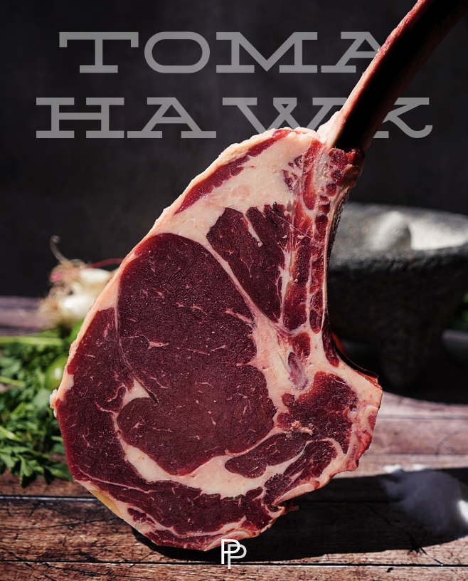 A close-up of a raw, marbled piece of beef with the bones attached, displayed on a wooden surface with herbs and garlic in the background.