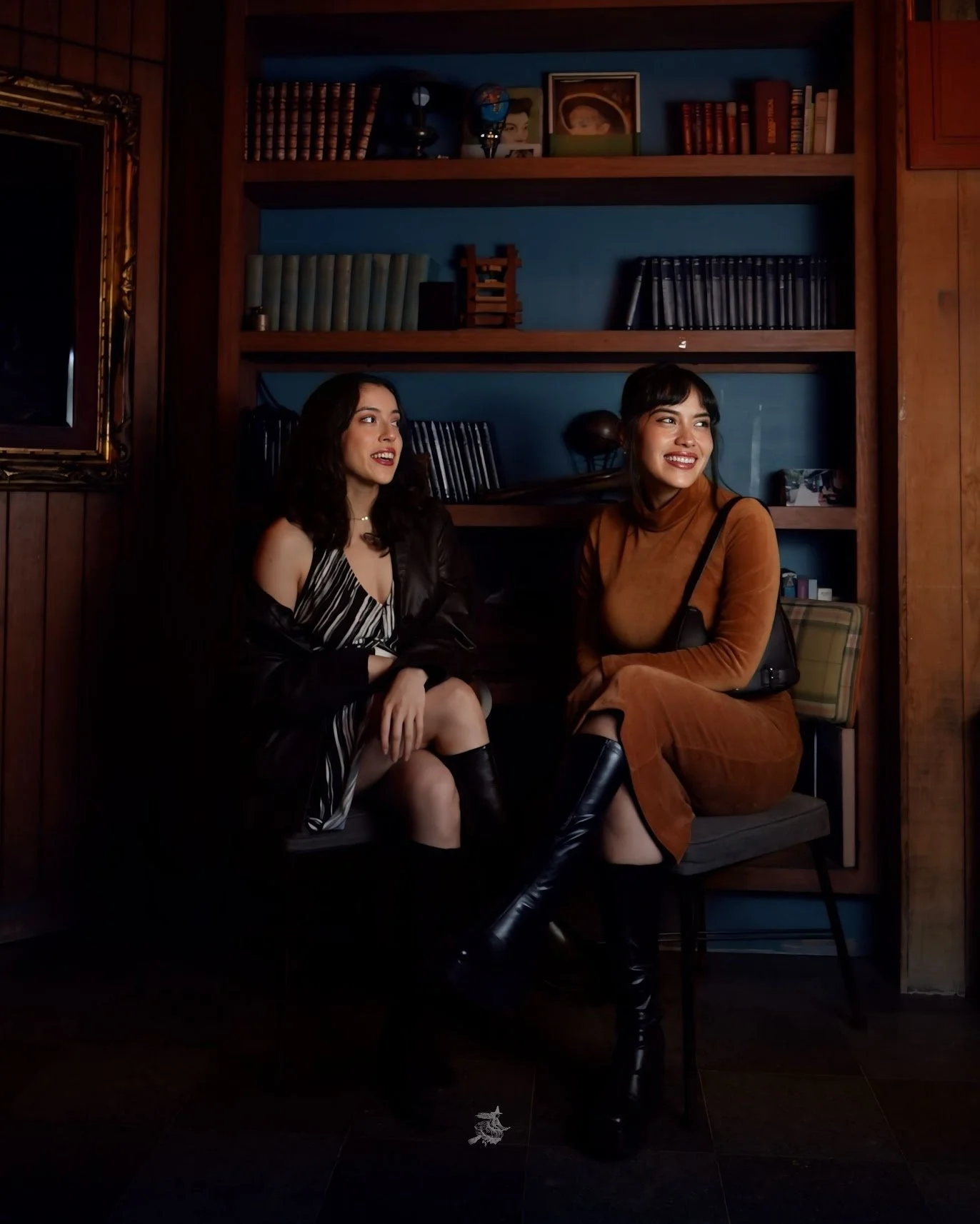 Two women sitting on a chair in a room with wooden walls and a bookshelf behind them. They are smiling and looking to their left. The woman on the left is wearing a black and white striped dress with a leather jacket, and the woman on the right is we