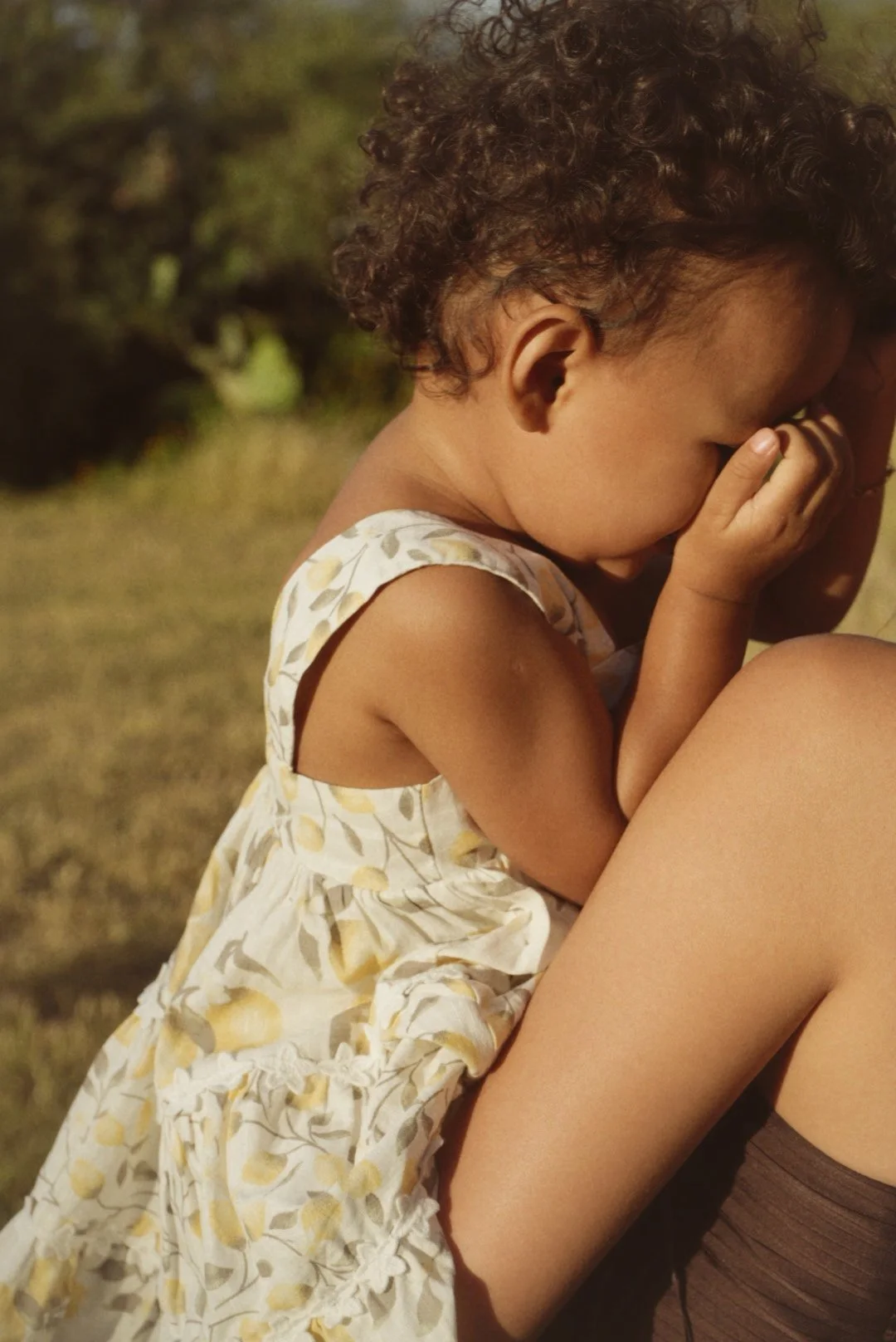 A young girl with curly hair is sitting outdoors, holding her face in her hands, with her eyes closed, and appears to be upset or crying.
