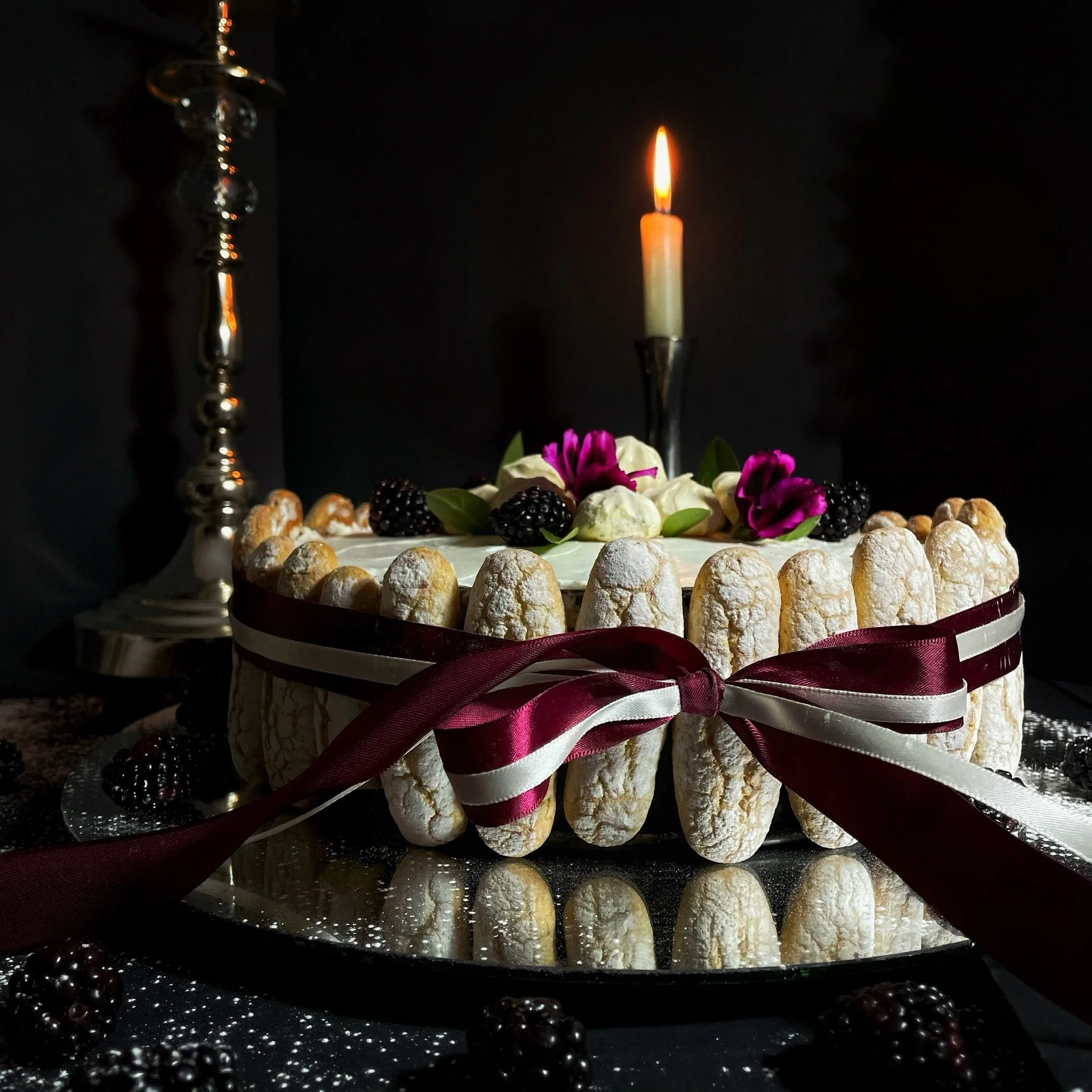 A cake decorated with blackberries, white and purple flowers, and a white candle with a lit flame, on a reflective surface. A tall candlestick is partially visible on the left side.