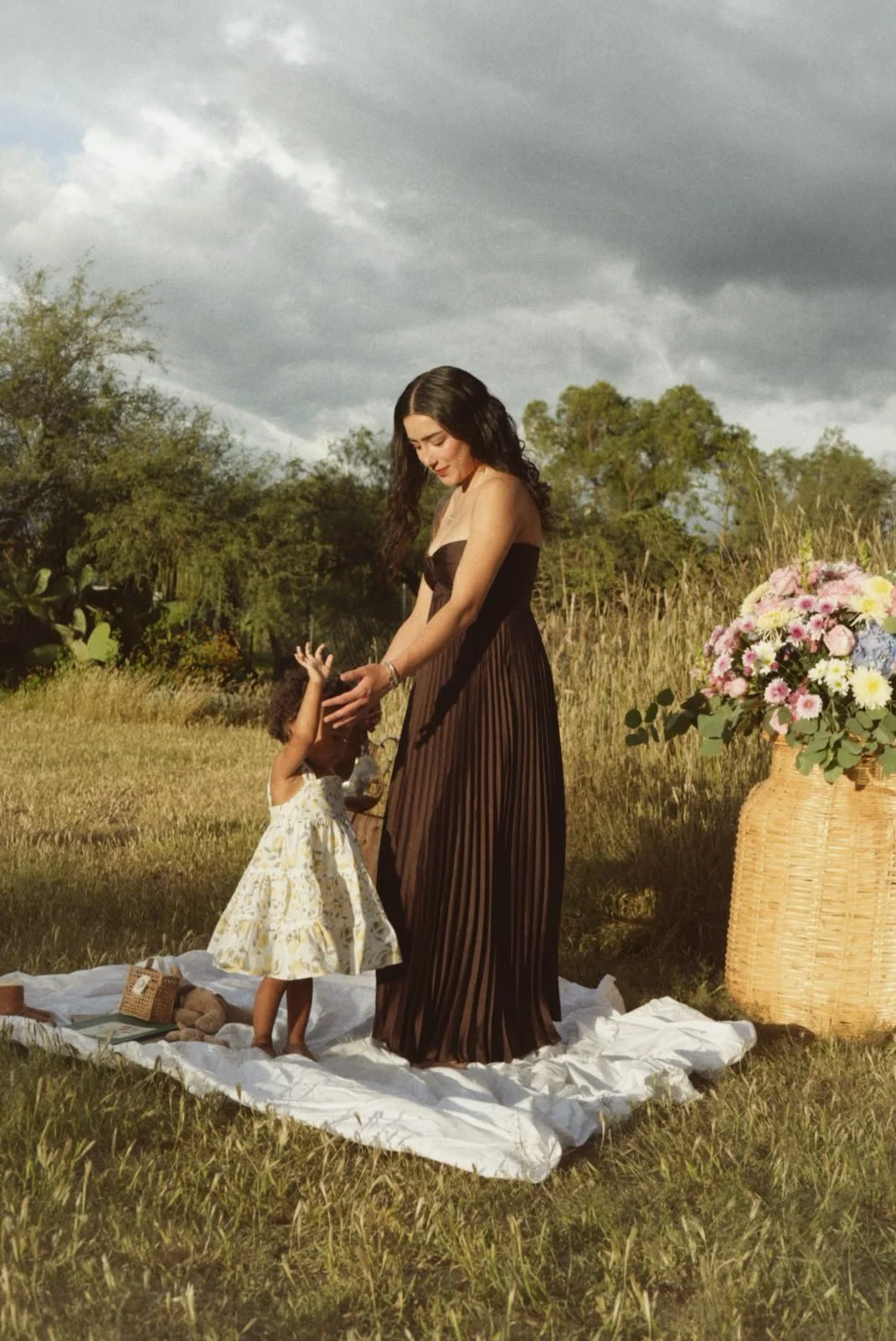 A woman in a brown pleated dress holding hands with a young girl in a white floral dress, standing on a white blanket outdoors in a field with trees and dark clouds in the sky.