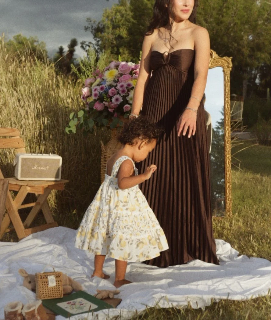 A woman in a strapless brown dress standing outdoors on a white sheet, with a young girl in a floral dress playing nearby. There is a bouquet of pink and white flowers, a vintage mirror, a small wooden table with a portable speaker, and toys on the g