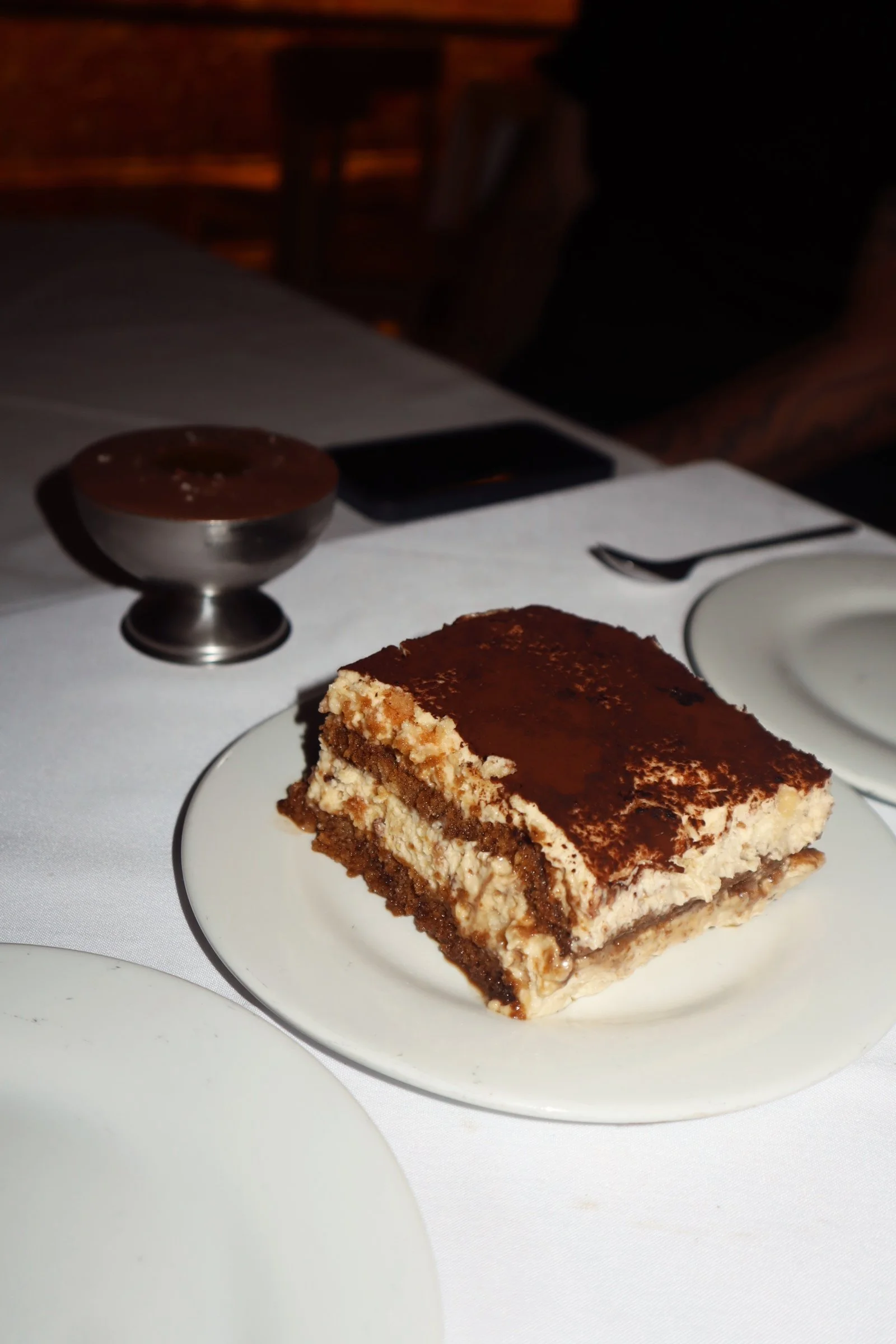 A slice of layered dessert with cream, chocolate, and cookie or biscuit layers on a white plate. A bowl of caramel sauce, a fork, and a chocolate bar are visible in the background on a white tablecloth.