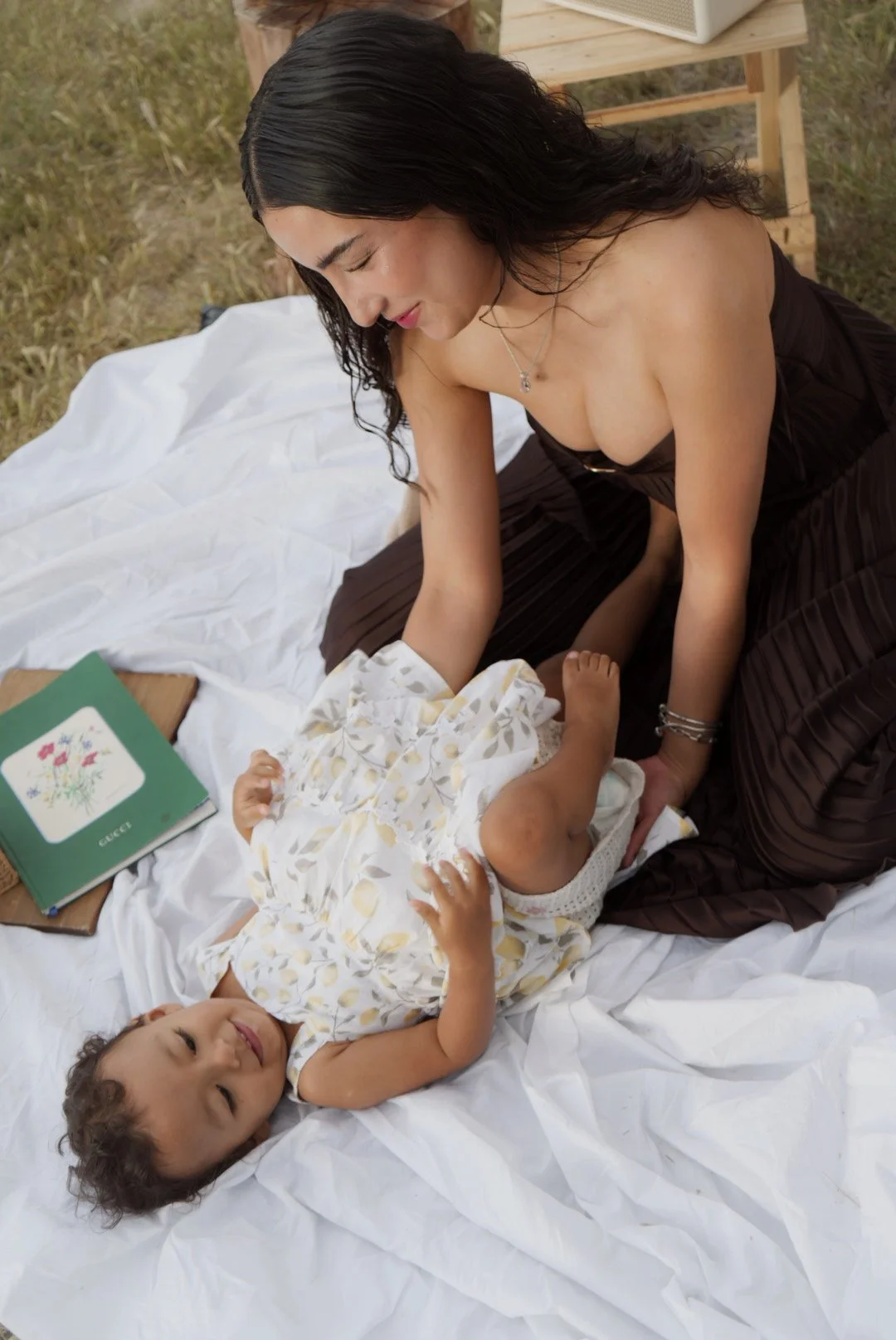 A woman with long dark hair, wearing a strapless black dress, sitting on a white sheet outdoors with two young children, one lying down and smiling at the camera, the other partially visible. The setting appears to be a picnic or outdoor photoshoot w