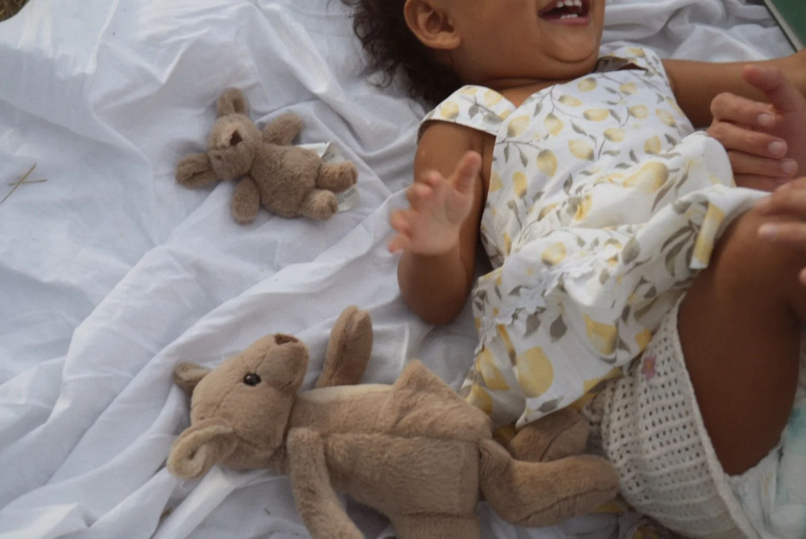 A smiling young child lying on a hospital bed, wearing a yellow and white patterned outfit, surrounded by plush teddy bears.