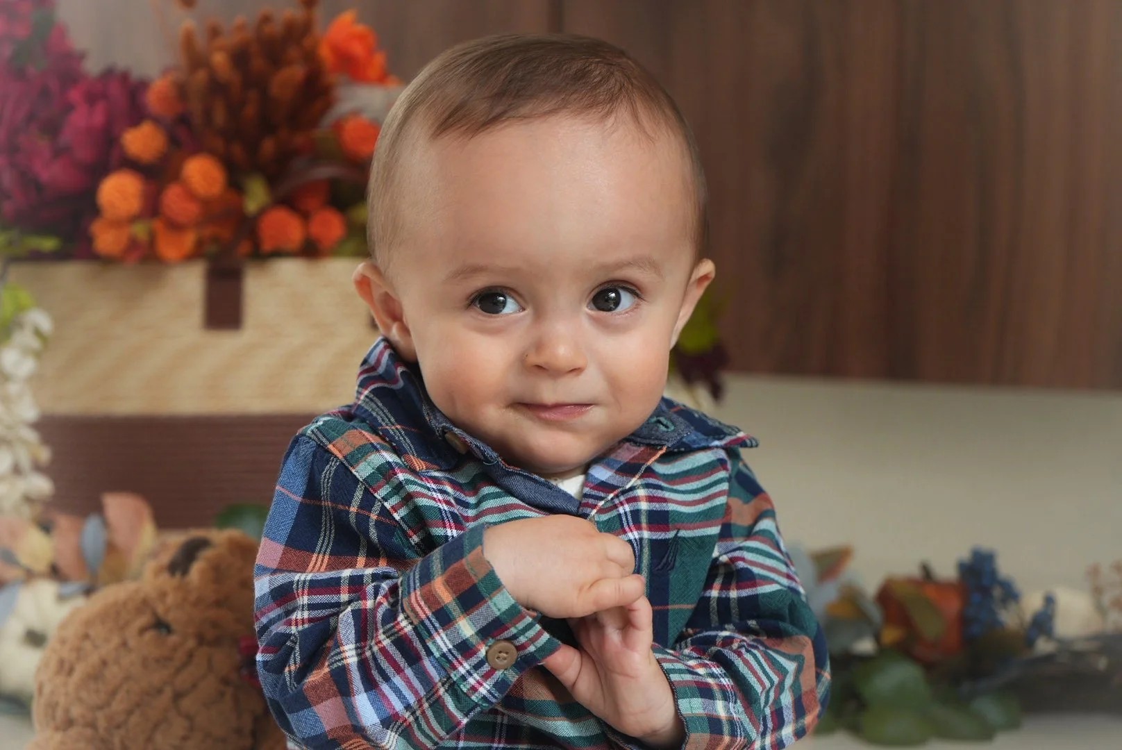 A young boy with big eyes and a slight smile wearing a plaid shirt, sitting indoors with a background of flowers and stuffed animals.