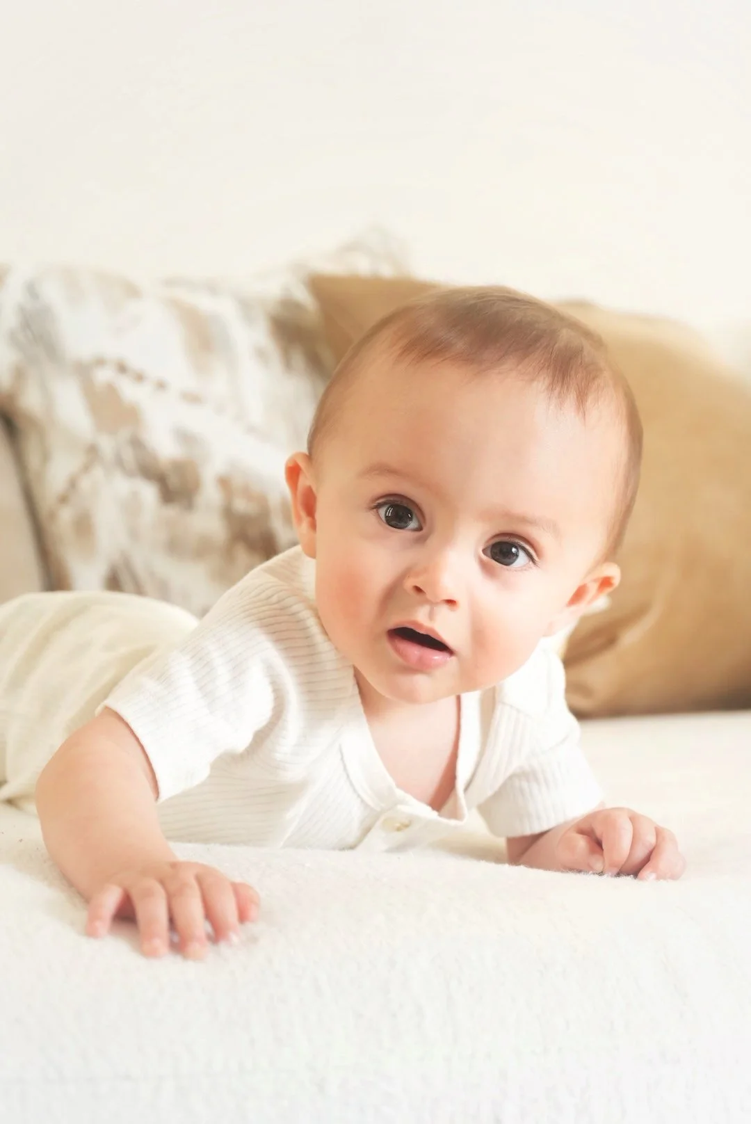 A baby boy with blue eyes and light brown hair lying on his stomach on a white bed, looking at the camera with a curious expression.