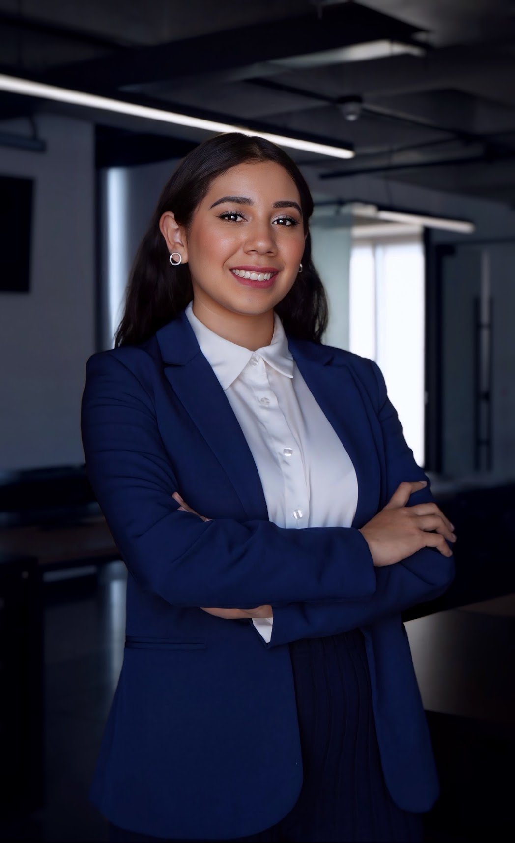 A young woman in a navy blazer and white shirt, standing with arms crossed in an office setting, smiling confidently.