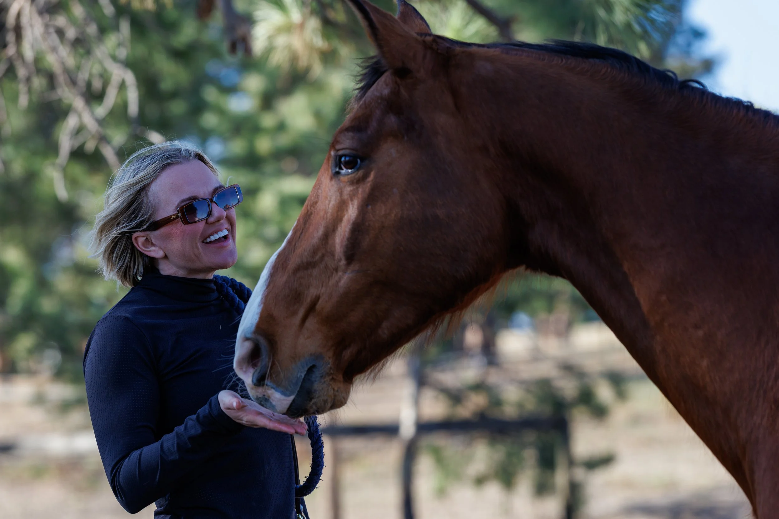 A woman wearing sunglasses and a black jacket smiling as a brown horse licks her hand in a natural outdoor setting with trees in the background.