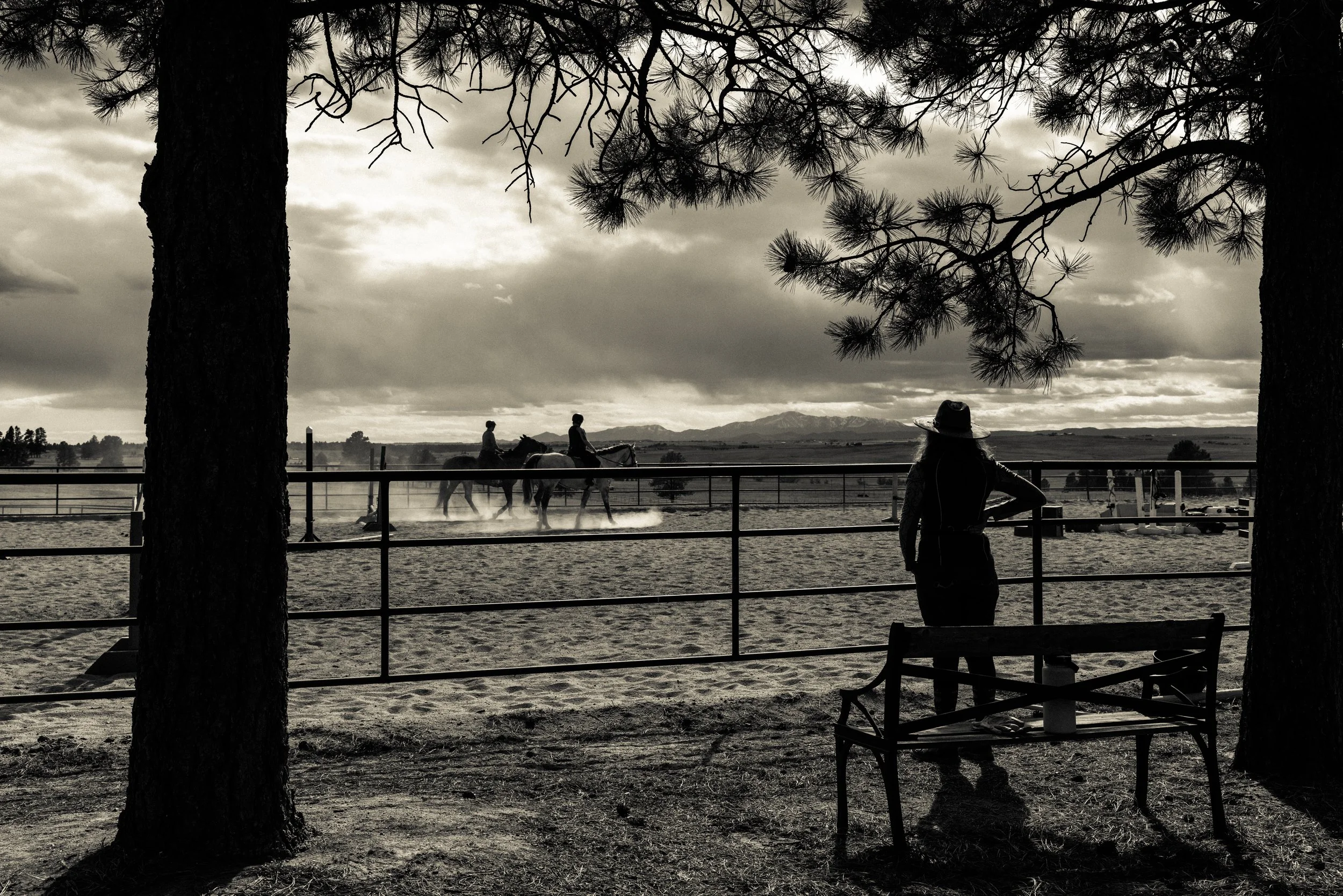 A woman standing behind a bench and a tree, watching two people riding horses in an outdoor arena, with mountains in the background and a cloudy sky, in a black-and-white photo.