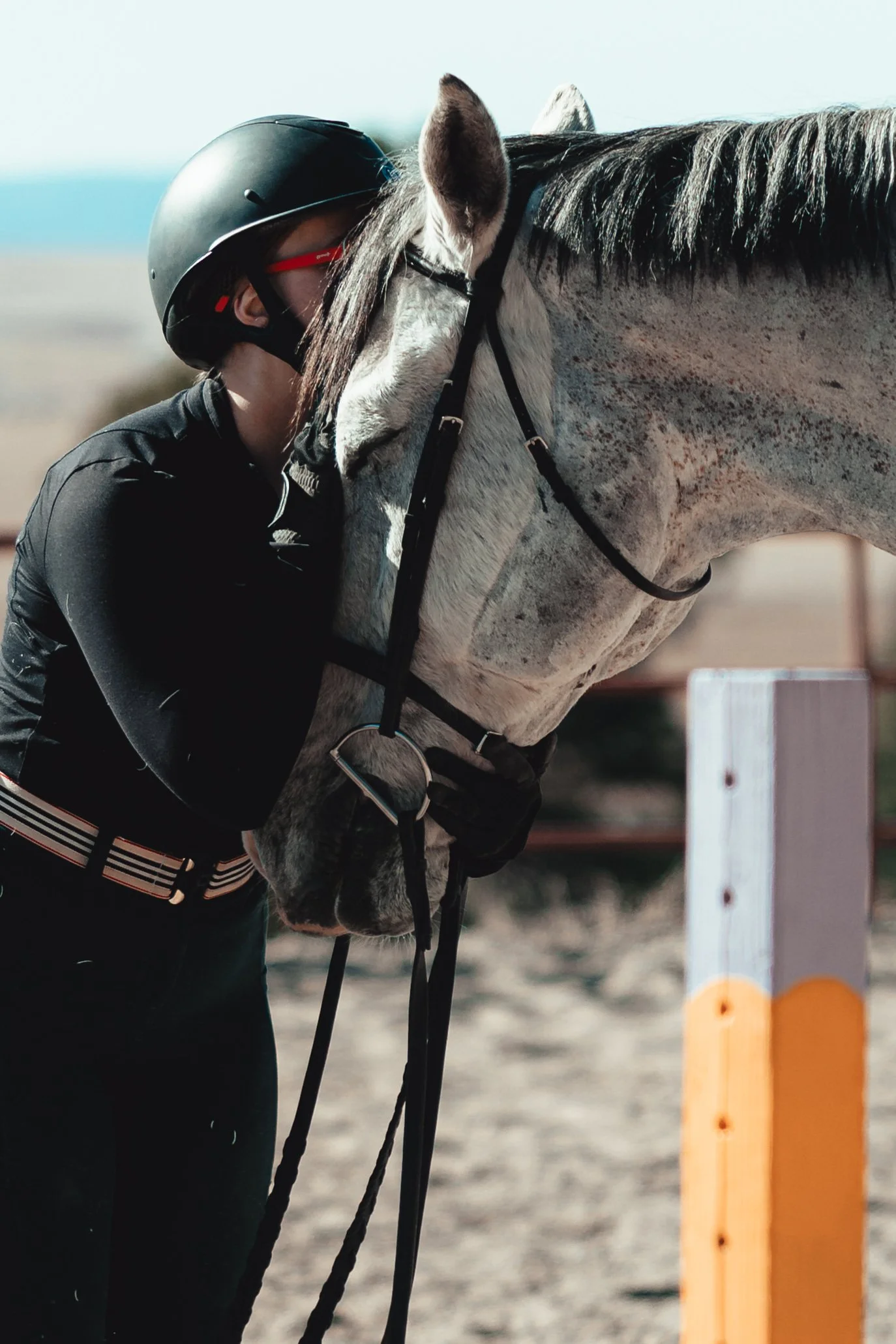 A person wearing a black riding helmet and black clothing leans in to kiss or nuzzle a white horse with gray spots, which has a black bridle, in an outdoor equestrian setting.